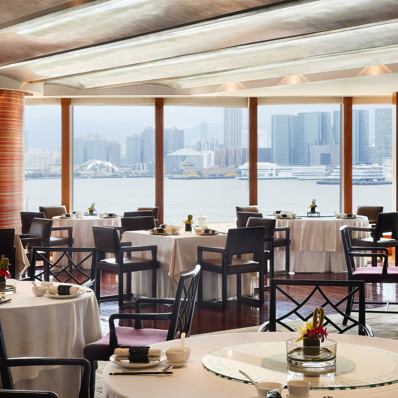 An elegant dining room with tables set with flowers and place settings, overlooking the city from across a body of water.