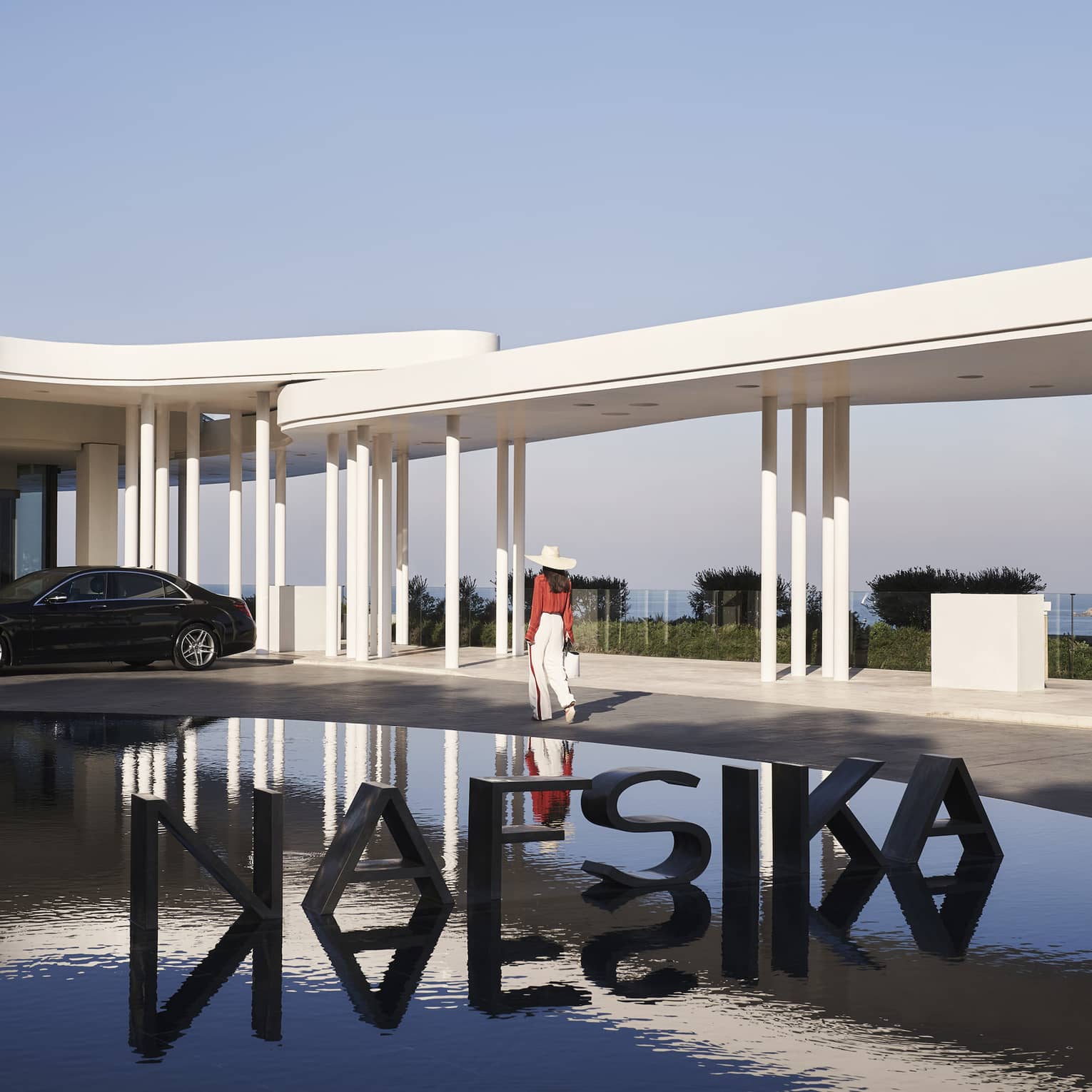 A guest passes a metal sign reading NAFSIKA in a fountain and towards an elegant car parked under modern white porticos.