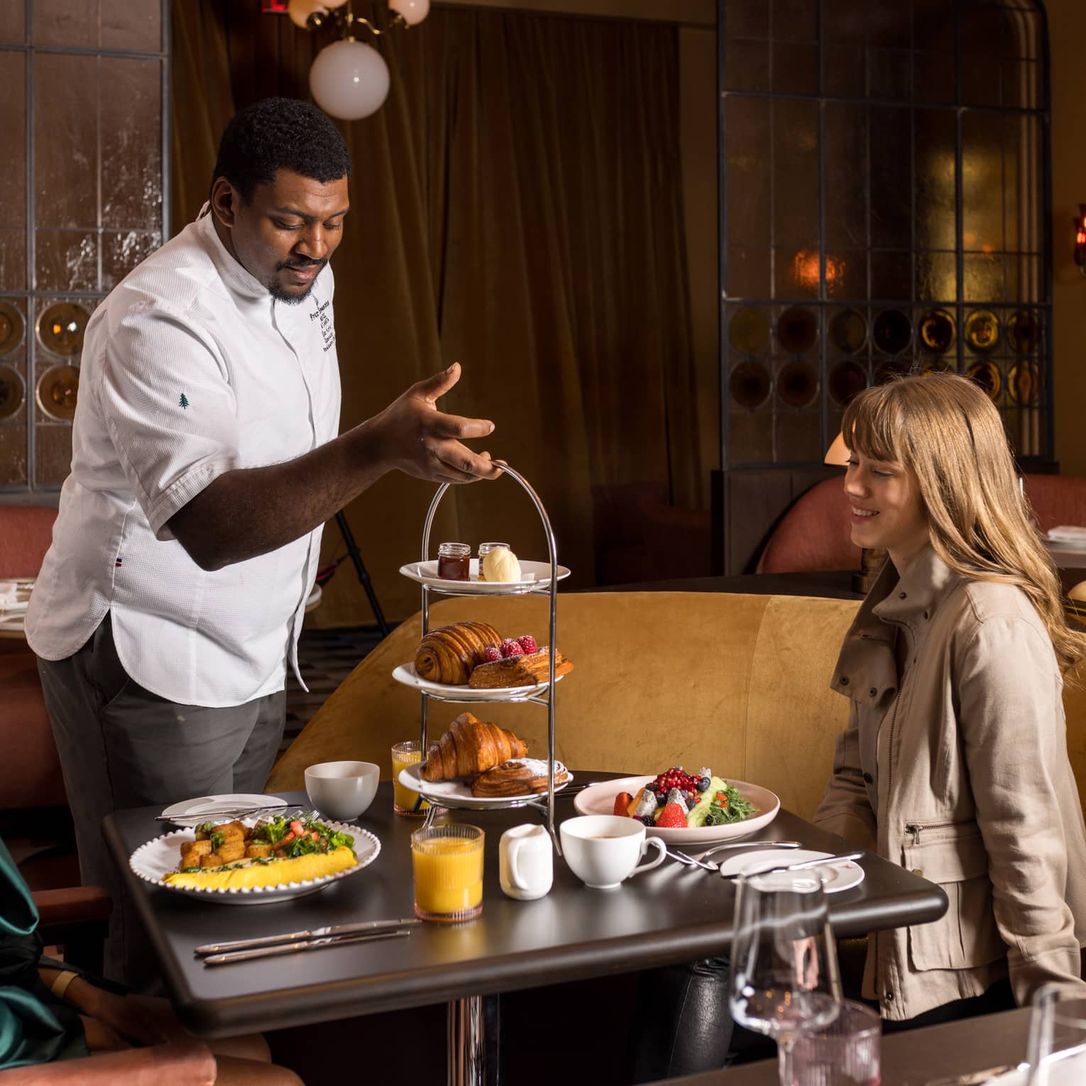 A person wearing a chef?s coat placing a three-tier display of sweets on a dining table