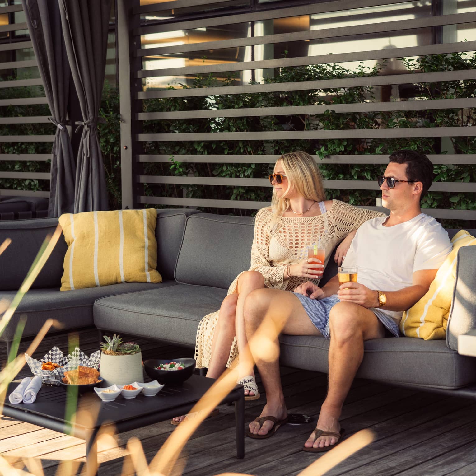 Two people sit on a patio couch with drinks, surrounded by cushions and snacks on a table. They wear sunglasses and casual summer attire.