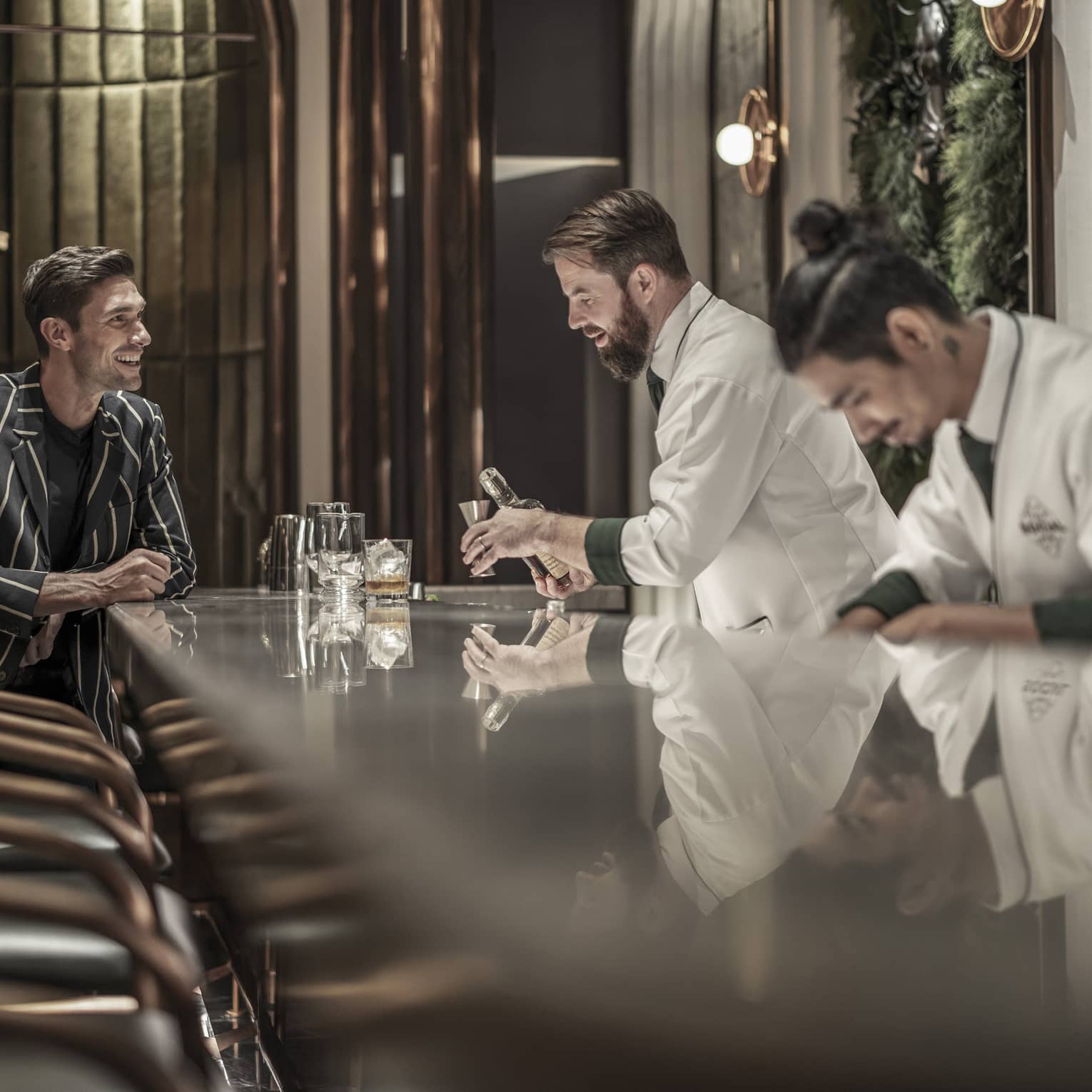 Bartender preparing a drink while a customer sits at the bar, both smiling, in a modern, upscale bar with a reflective countertop and warm lighting.