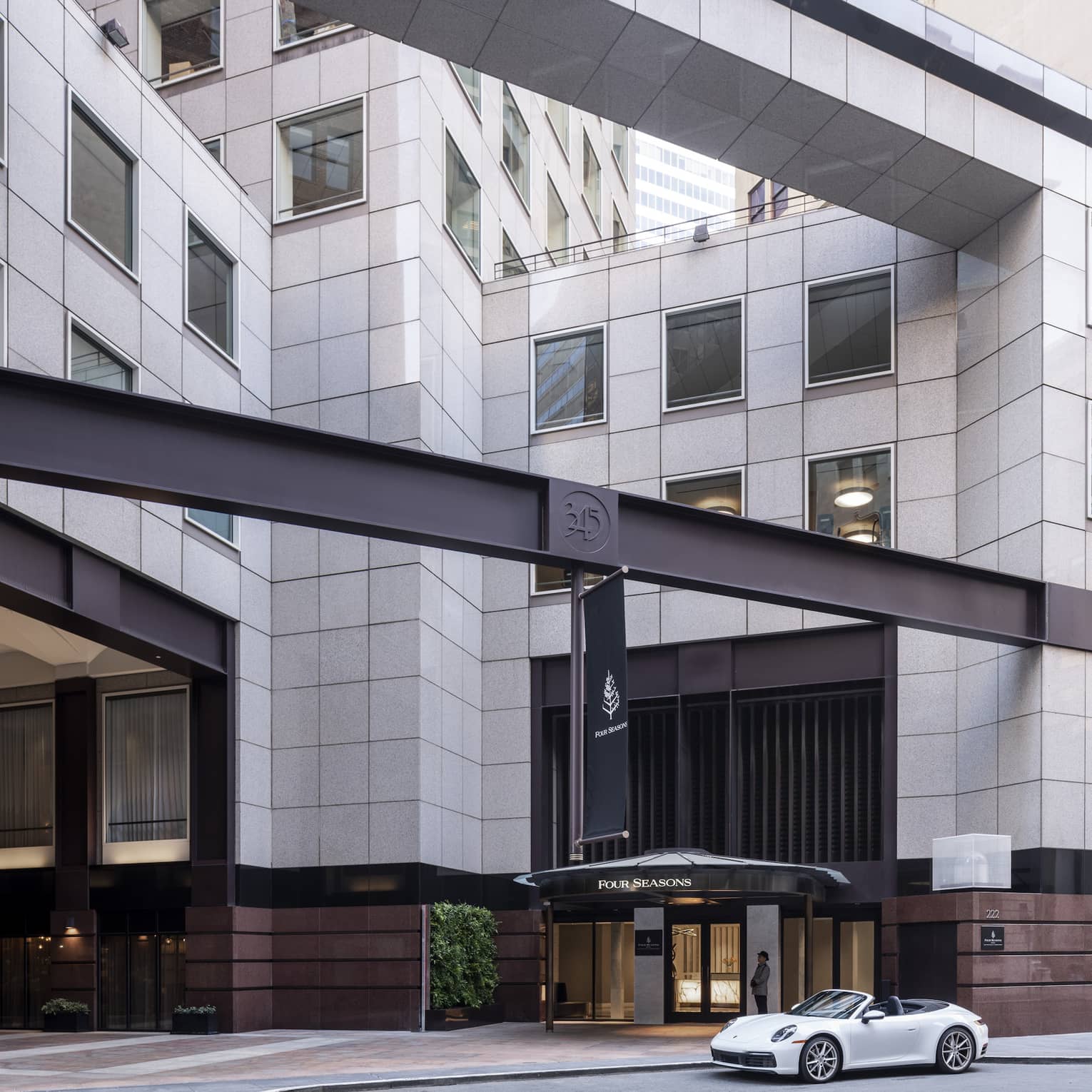 A white luxury sports caris parked in front of the massive Four Seasons hotel entrance, which is clad in pale grey granite.