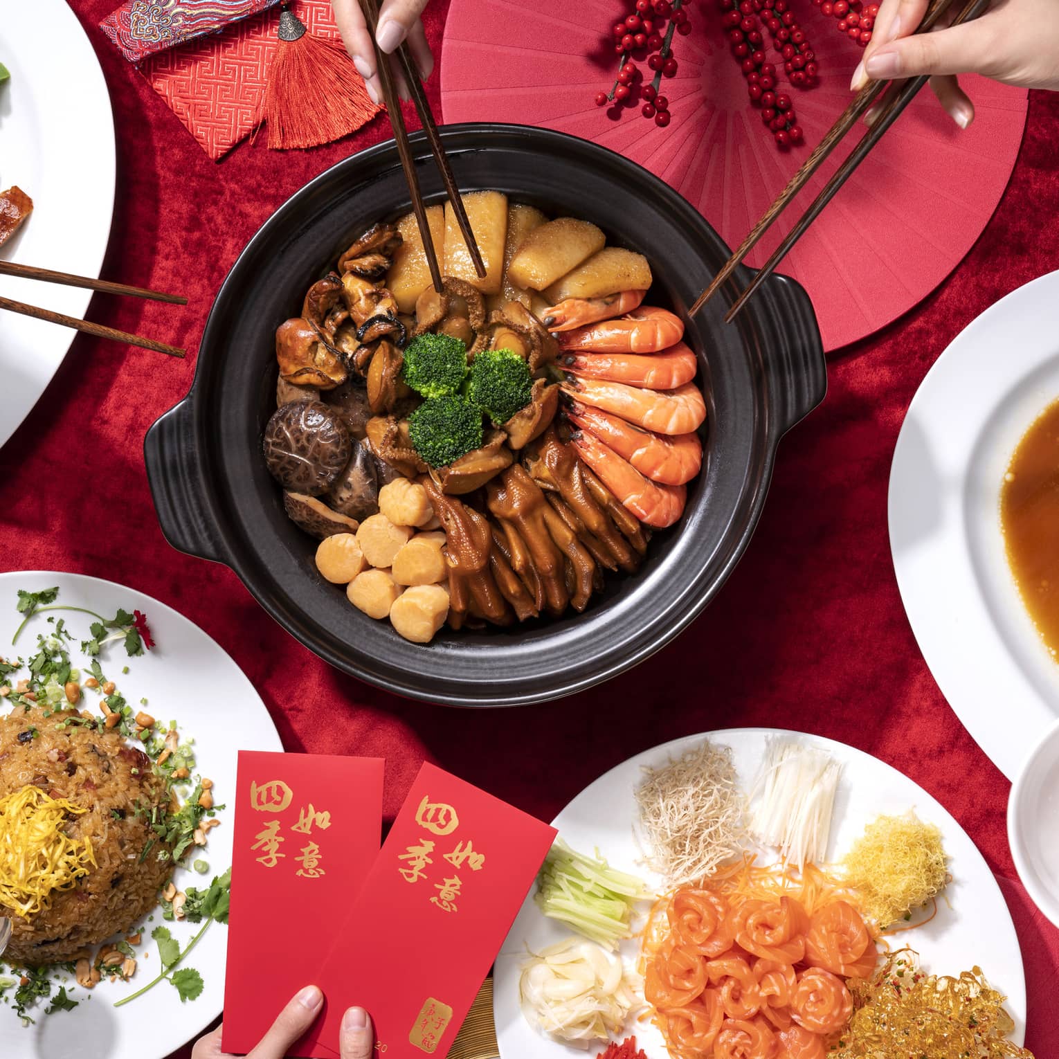 A festive Chinese New Year meal featuring a pot of delicacies, steamed fish, fried rice, smoked meats, and prosperity red packets on a red tablecloth.