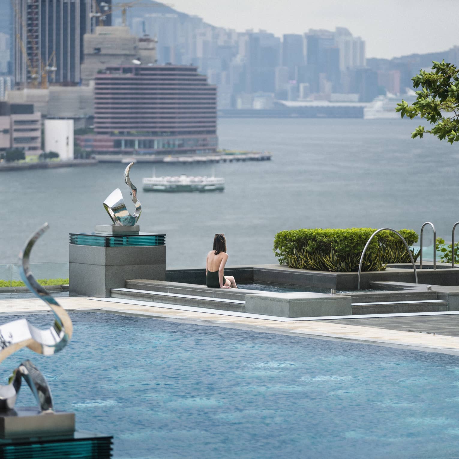 A person is seated at the edge of an outdoor swimming pool, which overlooks a large body of water with city buildings in the background.