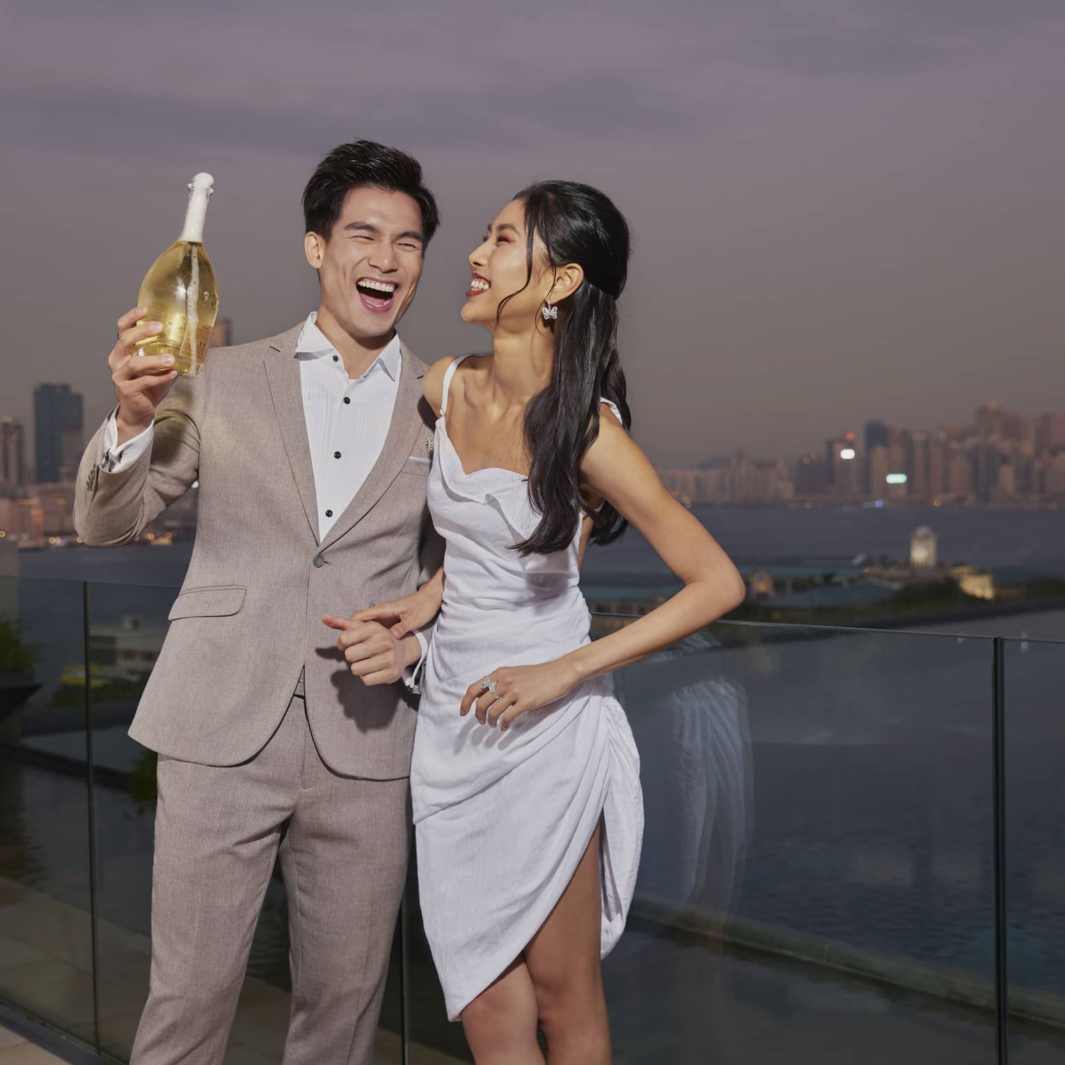 Man and woman laugh as they stand on outdoor pool terrace overlooking Victoria Harbor in Hong Kong, with bottle of sparkling wine in man's hand