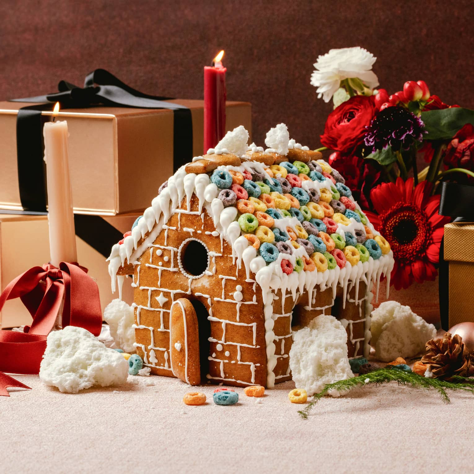 Gingerbread house decorated with icing and colourful cereal on the roof sits in front of three gold boxes wrapped in black ribbon, two pillar candles and a bouquet of red and white flowers
