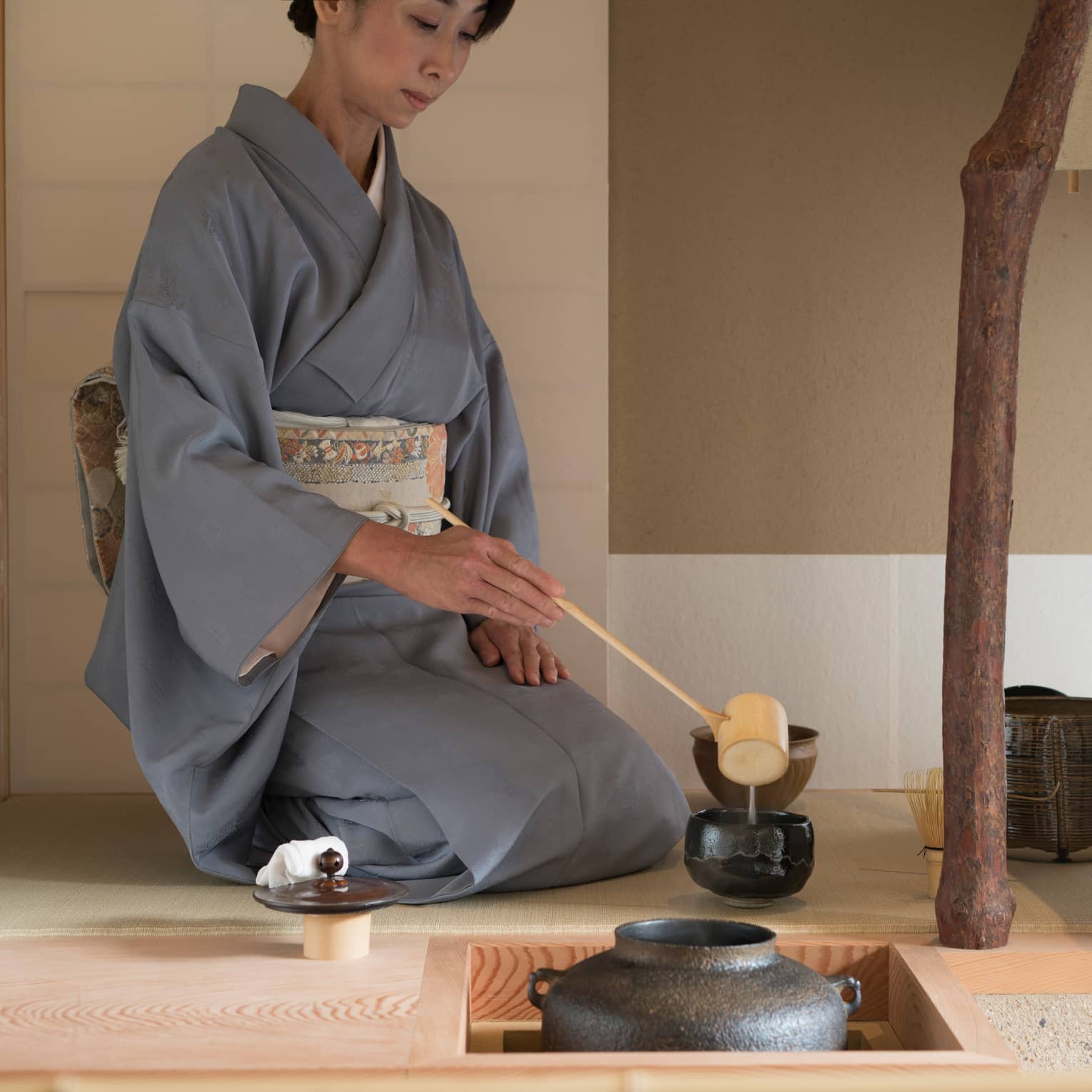 Woman in robe kneels on floor, ladles tea into cup with wood spoon