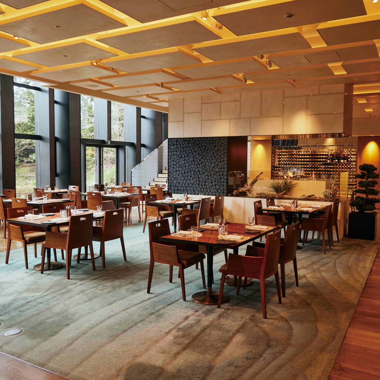 Restaurant dining room with warm lighting, light wood floors, floor-to-ceiling windows and green patterned area rug