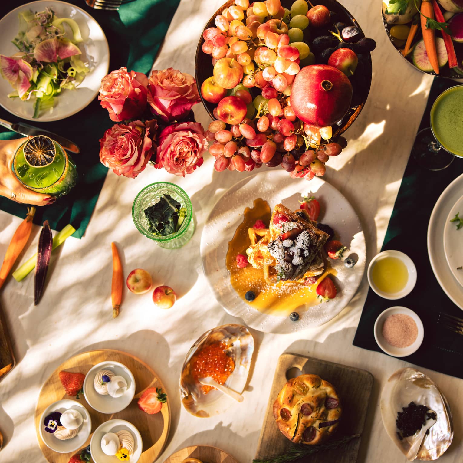 Overhead view of a sun-dappled white marble dining table filled with brunch dishes, fresh fruit and cocktails