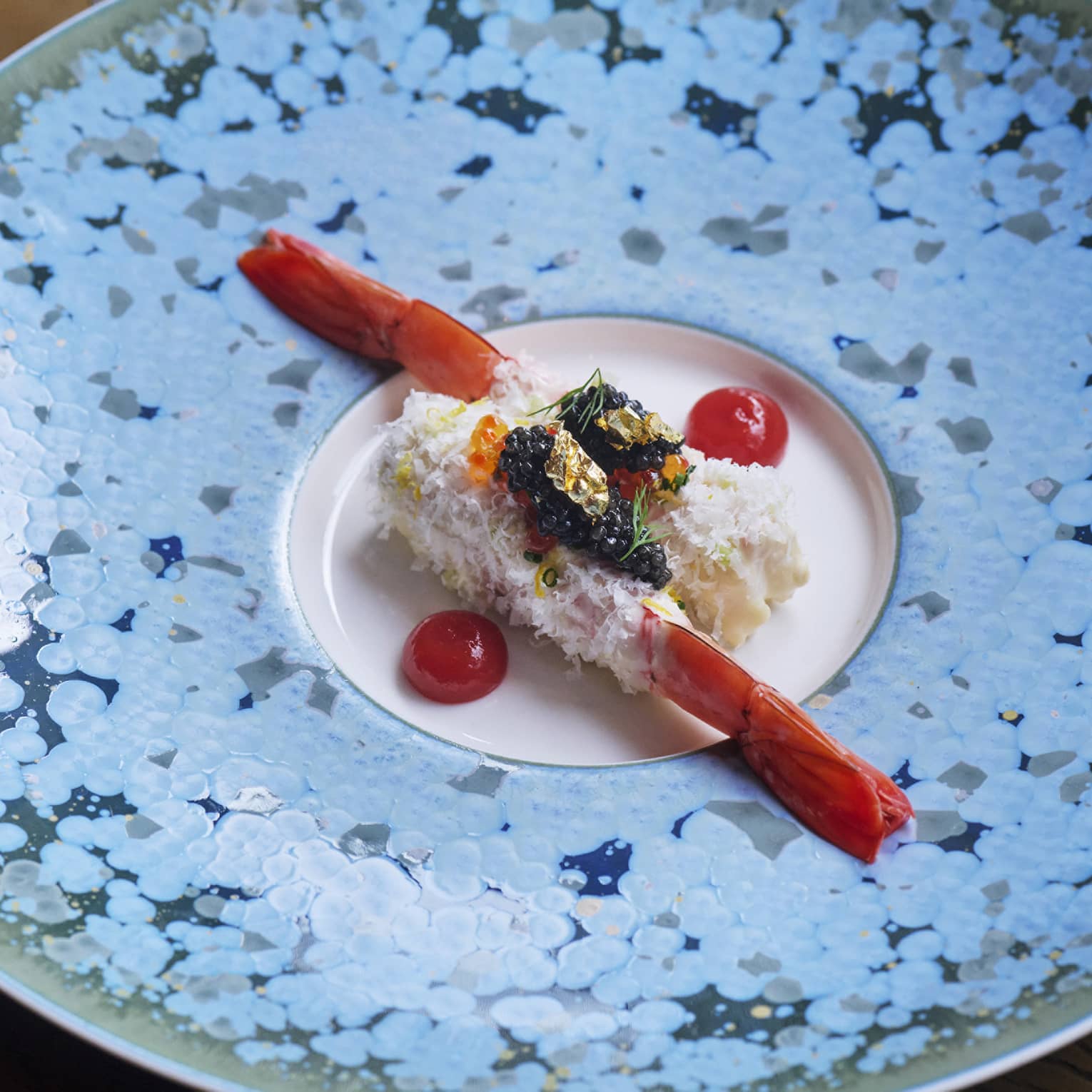 Two elegantly plated prawns topped with garnishes and flanked by cherry tomato halves in centre of blue speckled plate