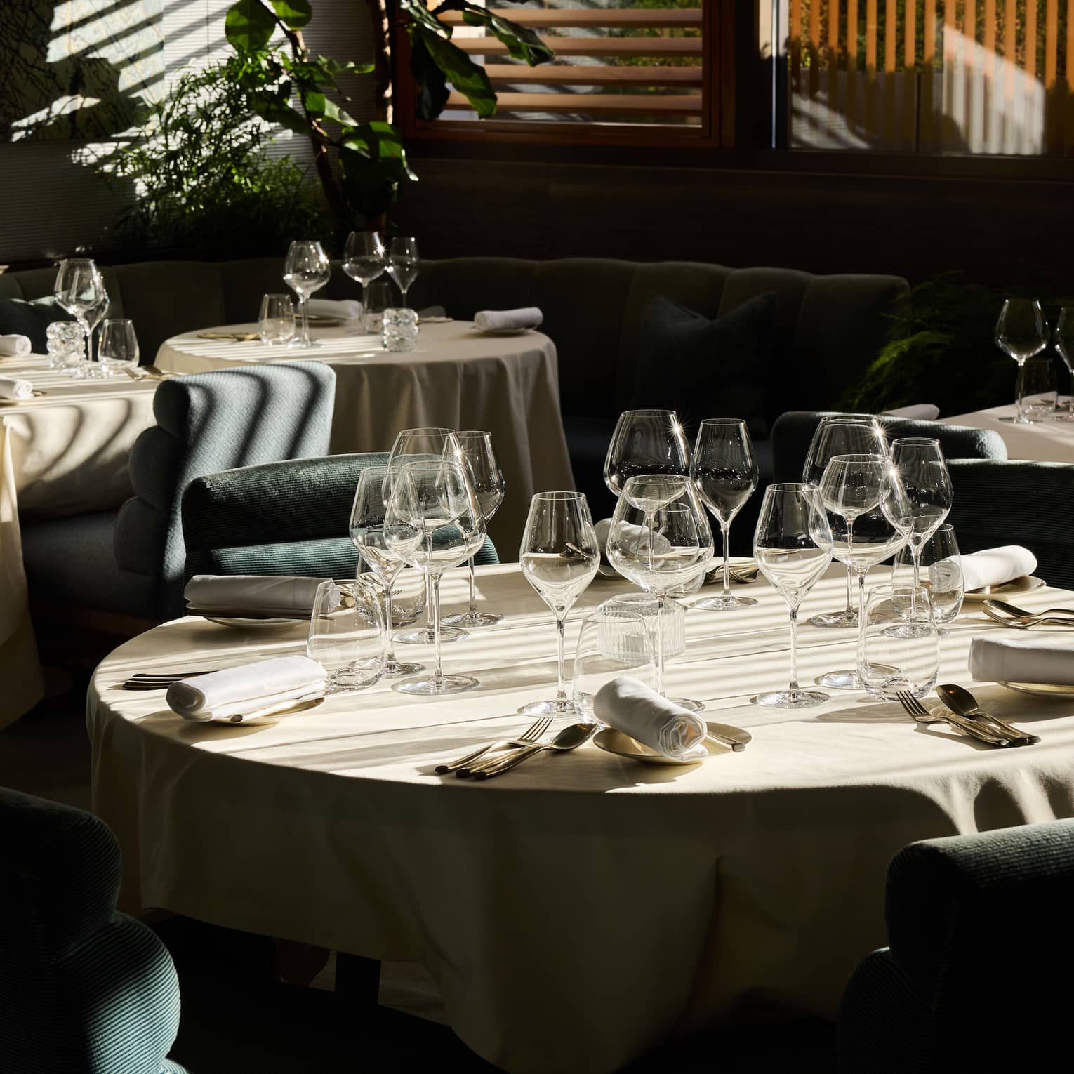 An indoor dining space with dramatic daylight filtering through window blinds and featuring tables, chairs, and place settings for a meal