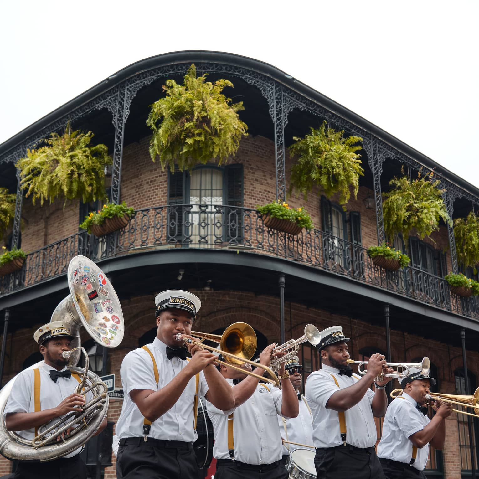 Six-piece brass band plays at the corner of a two-storey building beneath a balcony adorned with flags, ferns and flowers
