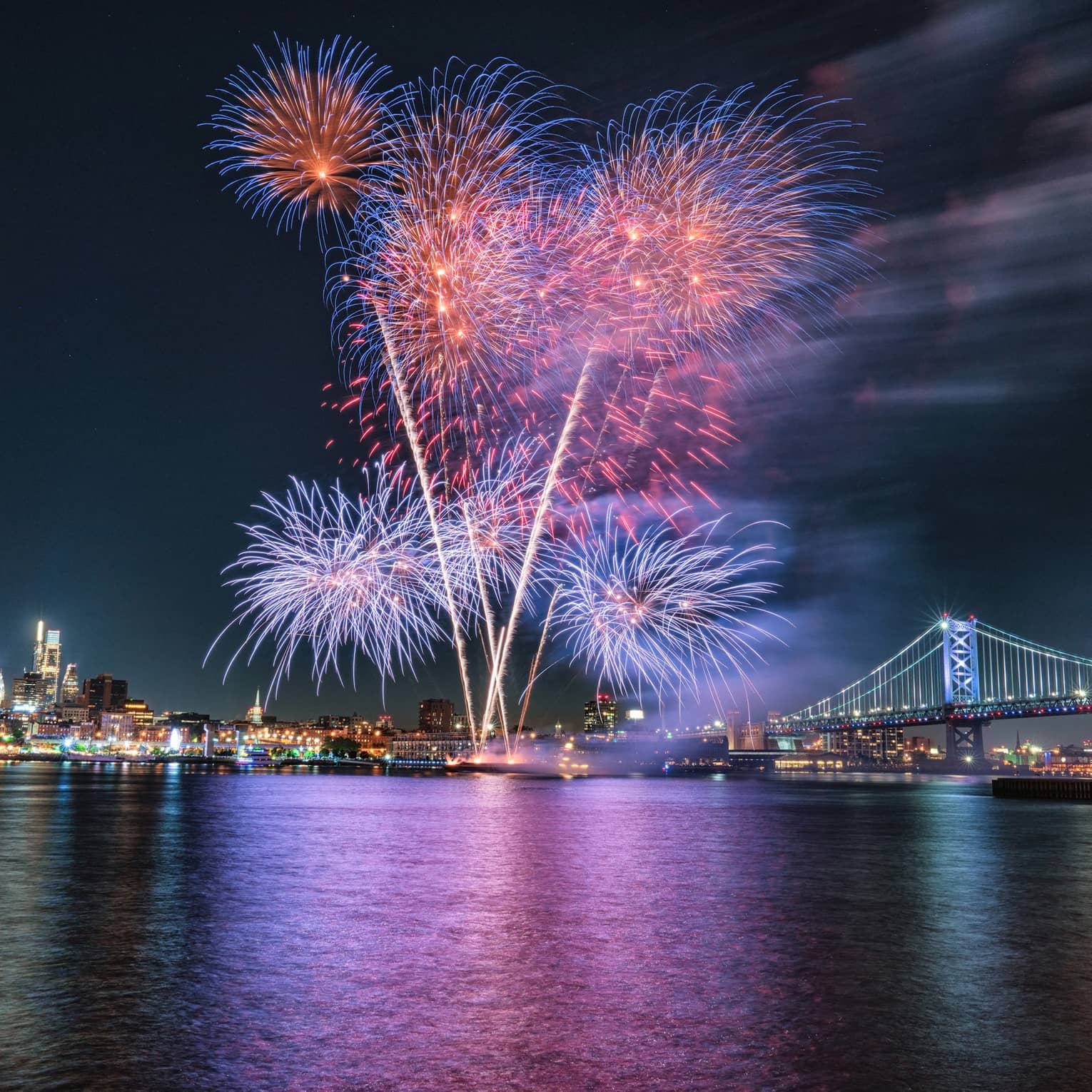 Purple, pink and blue fireworks exploding in the night sky above a city and a large metal bridge.