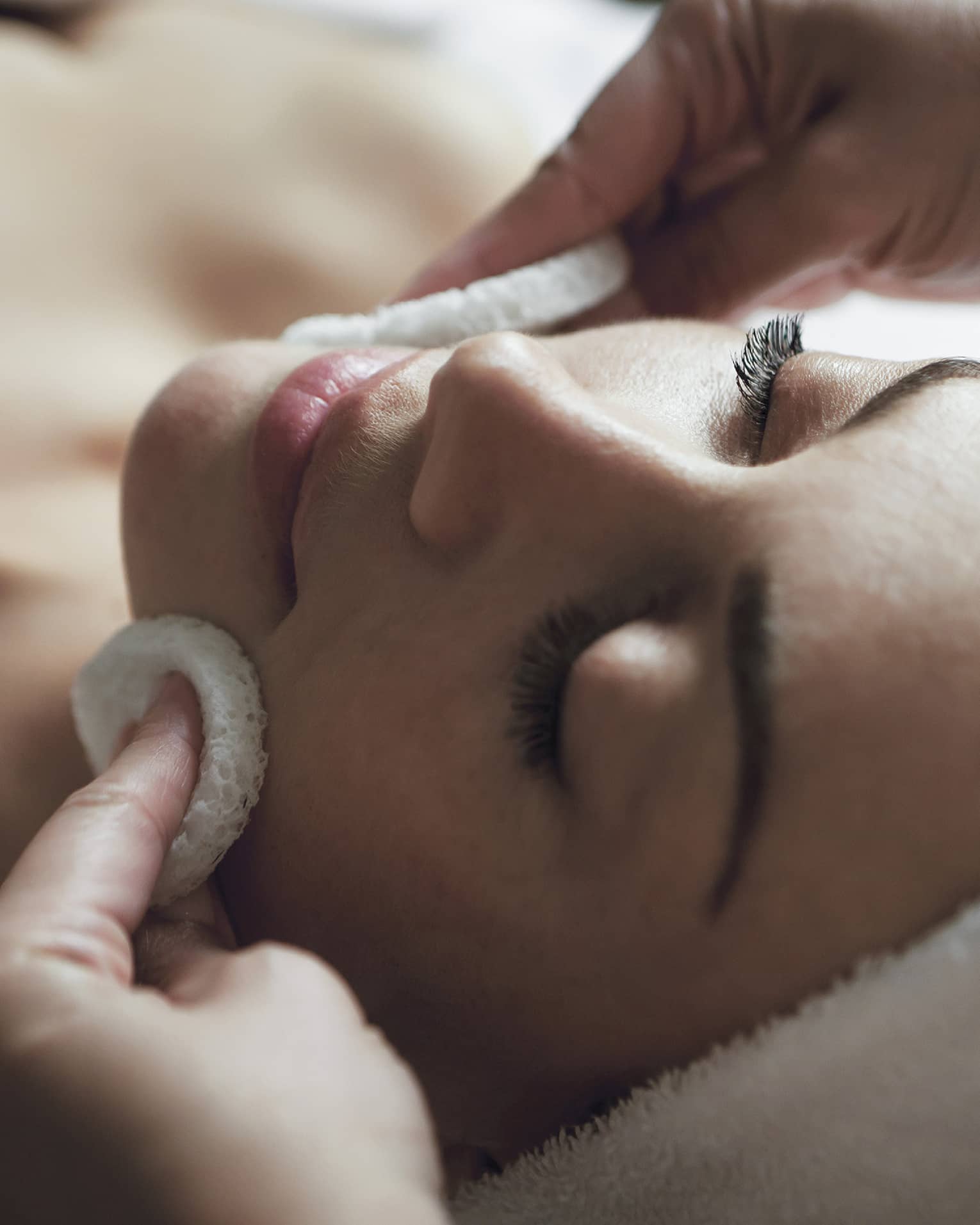 A detail of hands rubbing a sponge on a woman's face as she lies on a massage table with her eyes closed and hair wrapped up in a white towel
