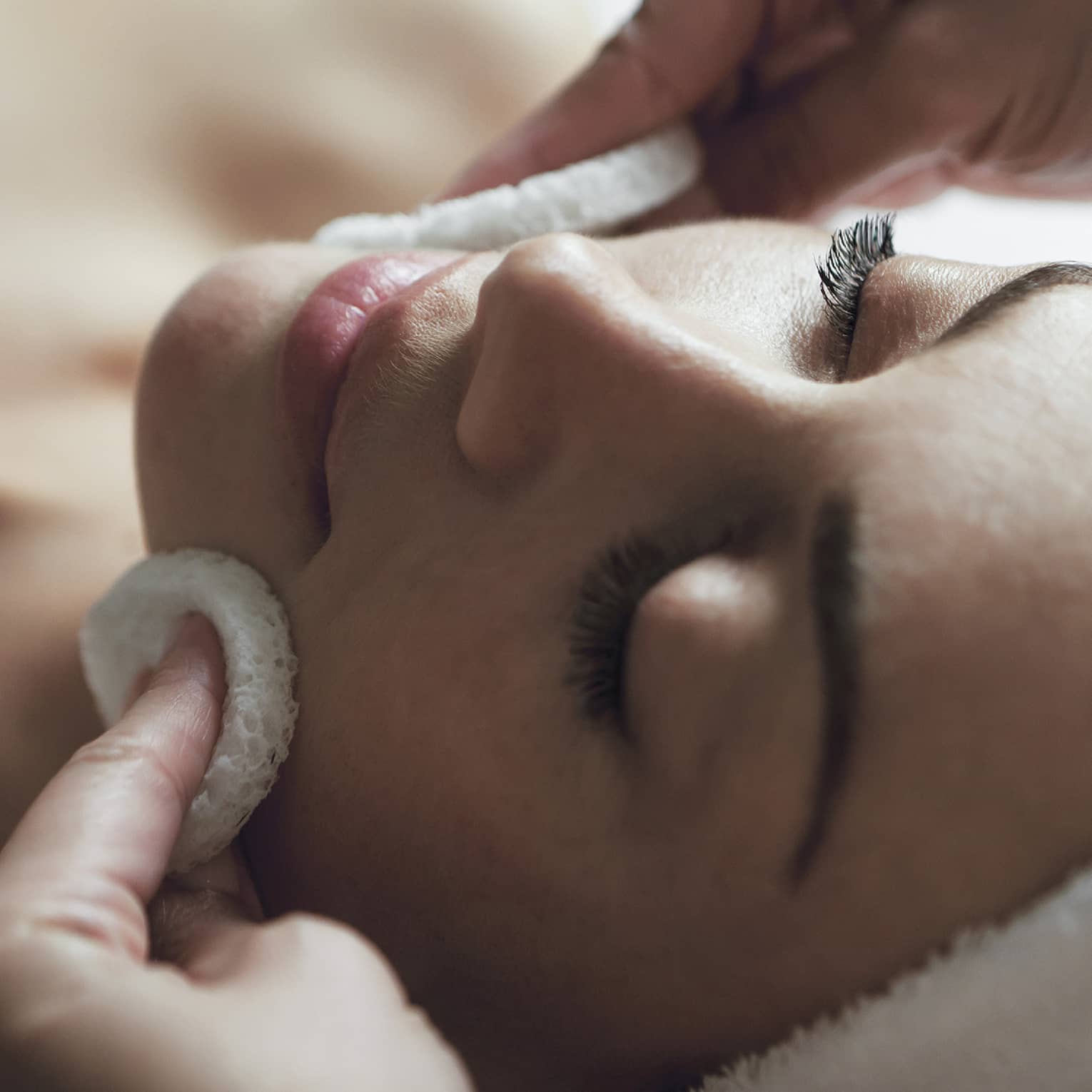 A detail of hands rubbing a sponge on a woman's face as she lies on a massage table with her eyes closed and hair wrapped up in a white towel