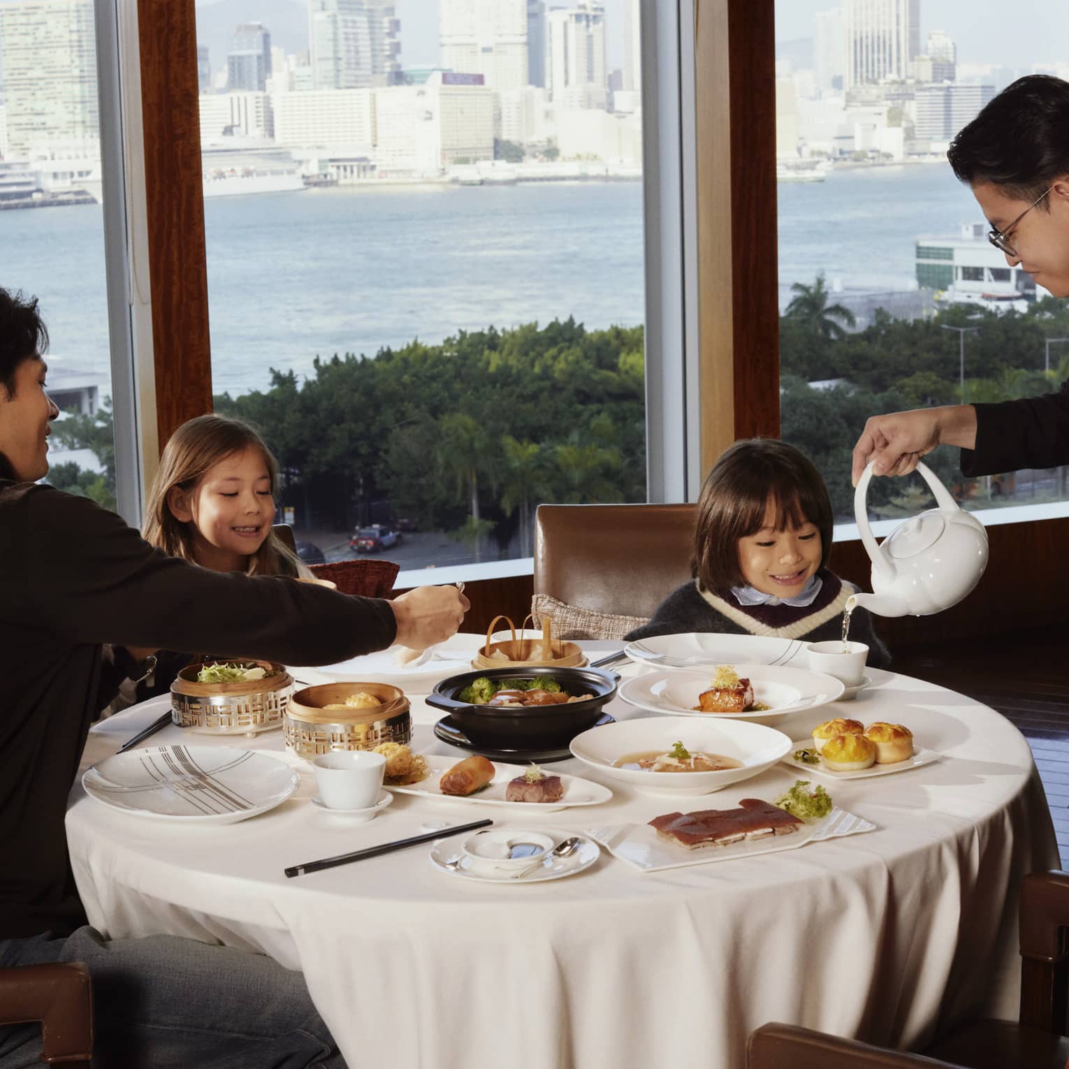 A hotel restaurant waiter pour tea at a white tablecloth table with man and two children, set next to a window overlooking city and harbour