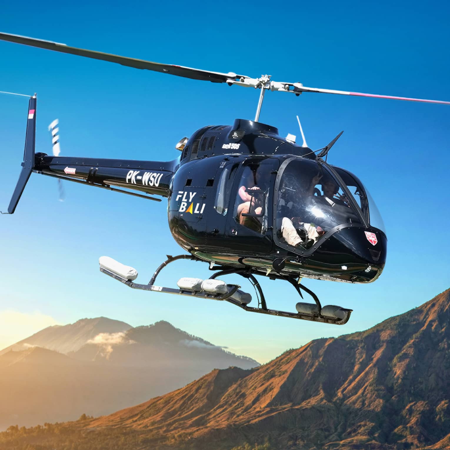 A helicopter glides across a clear blue sky past tree-lined mountains partially covered by low-lying clouds.