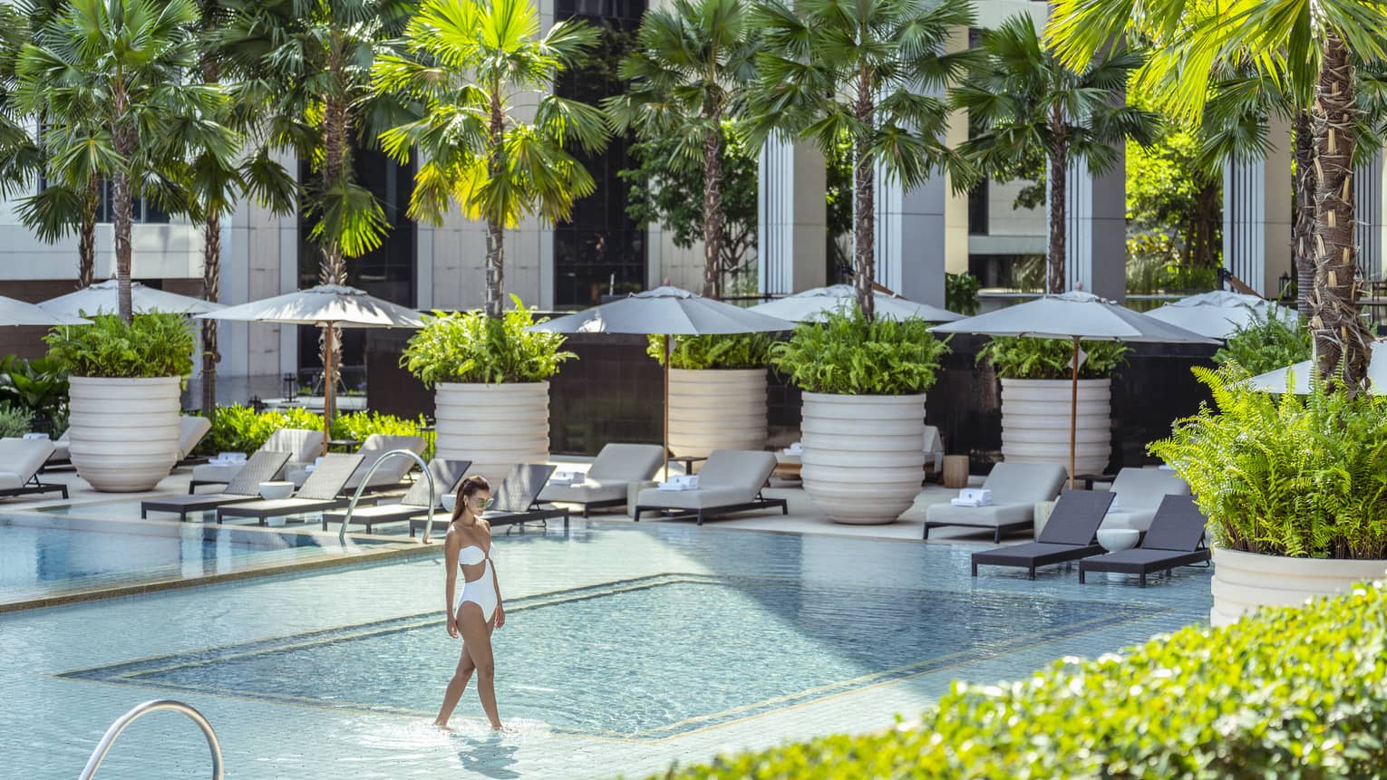 Woman in white bathing suit wades in outdoor pool surrounded by lounge chairs with umbrellas and palm trees