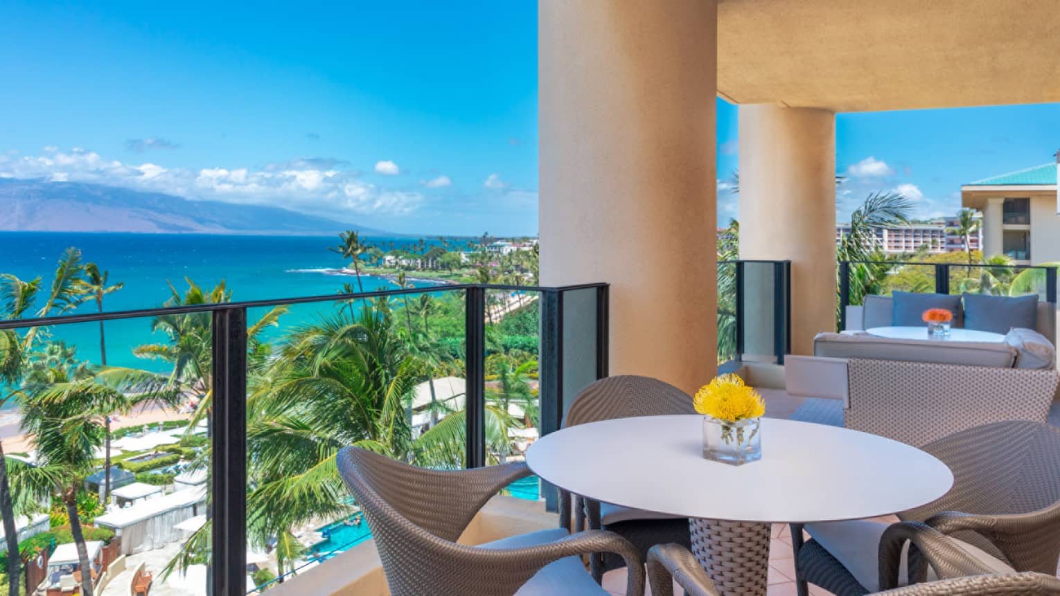 Luxury resort balcony with dining table and seating area, overlooking ocean, palm trees and mountain views