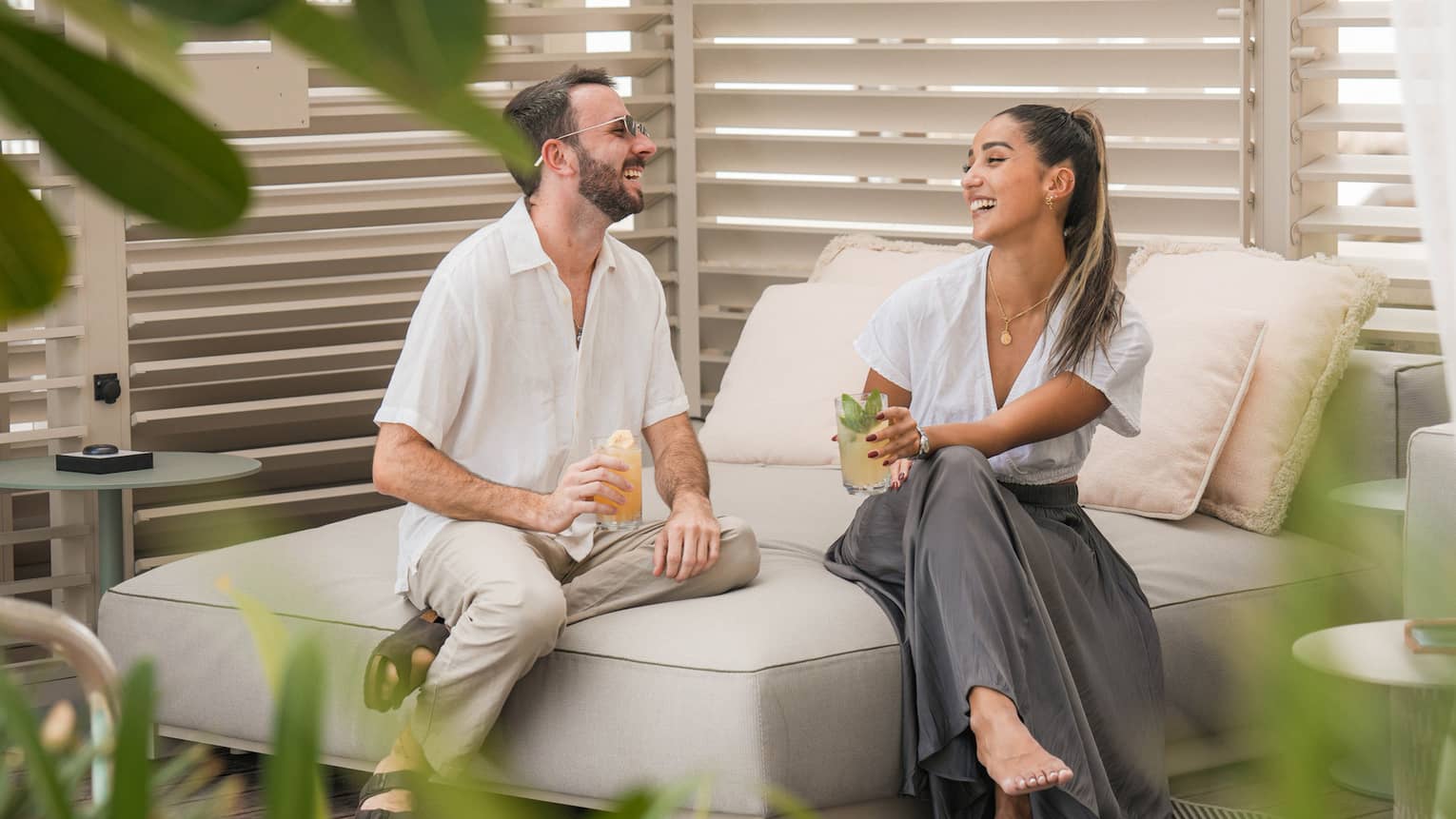 Two guests enjoy iced cocktails in a private cabana decorated with shuttered windows, leafy ferns and a plush chaise.
