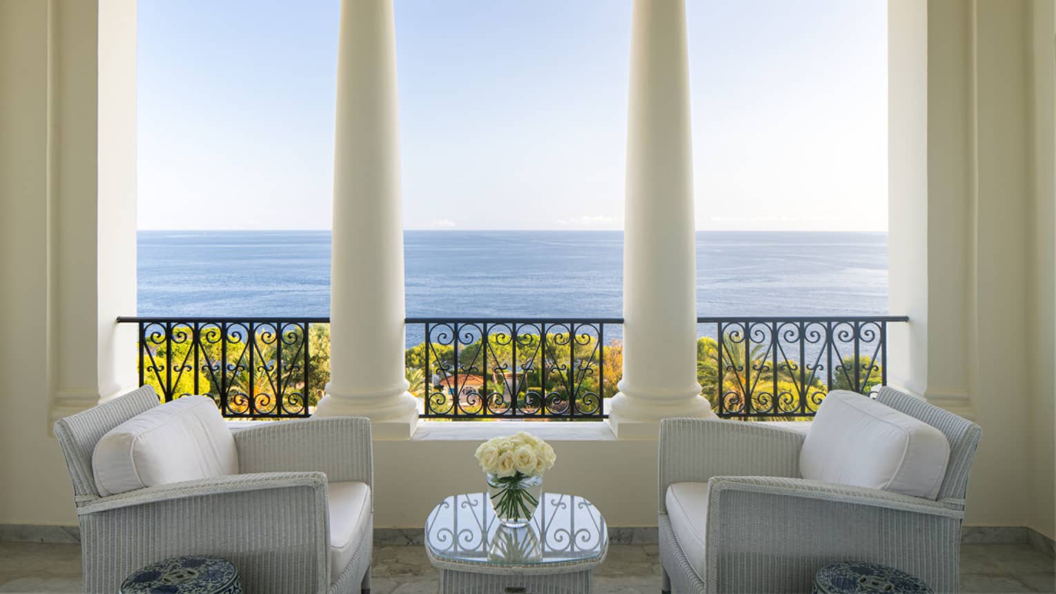 Two white club chairs facing with table between and beside white columns and terrace with sea view