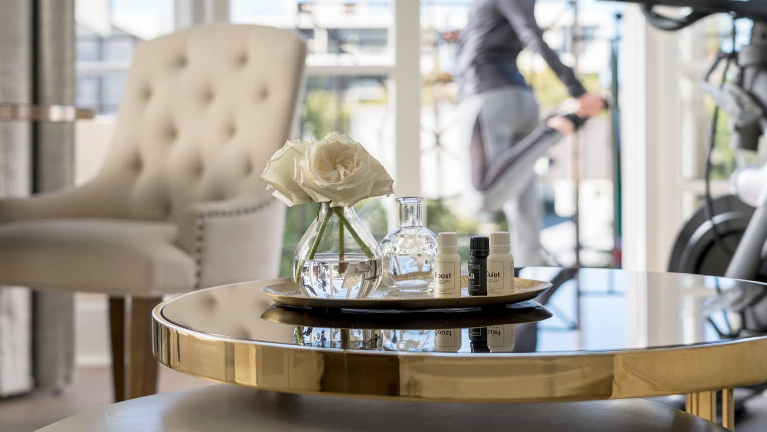 Spa tray with fresh white flowers on table by treadmill, woman stretching at balcony door