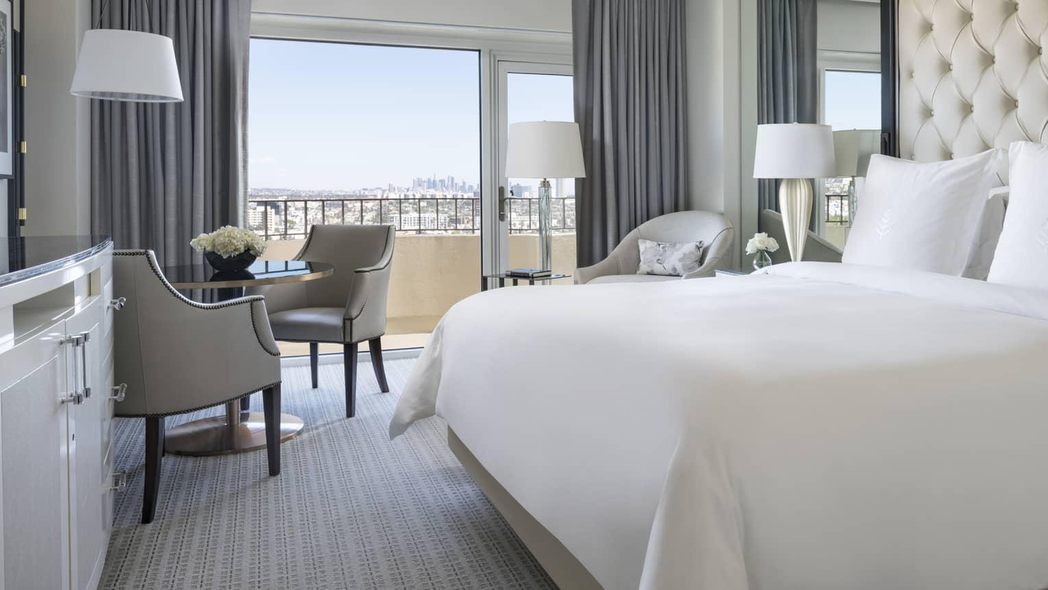 Premier Balcony Room hotel bed against padded, tufted wall, mirror, cafe table by sunny window