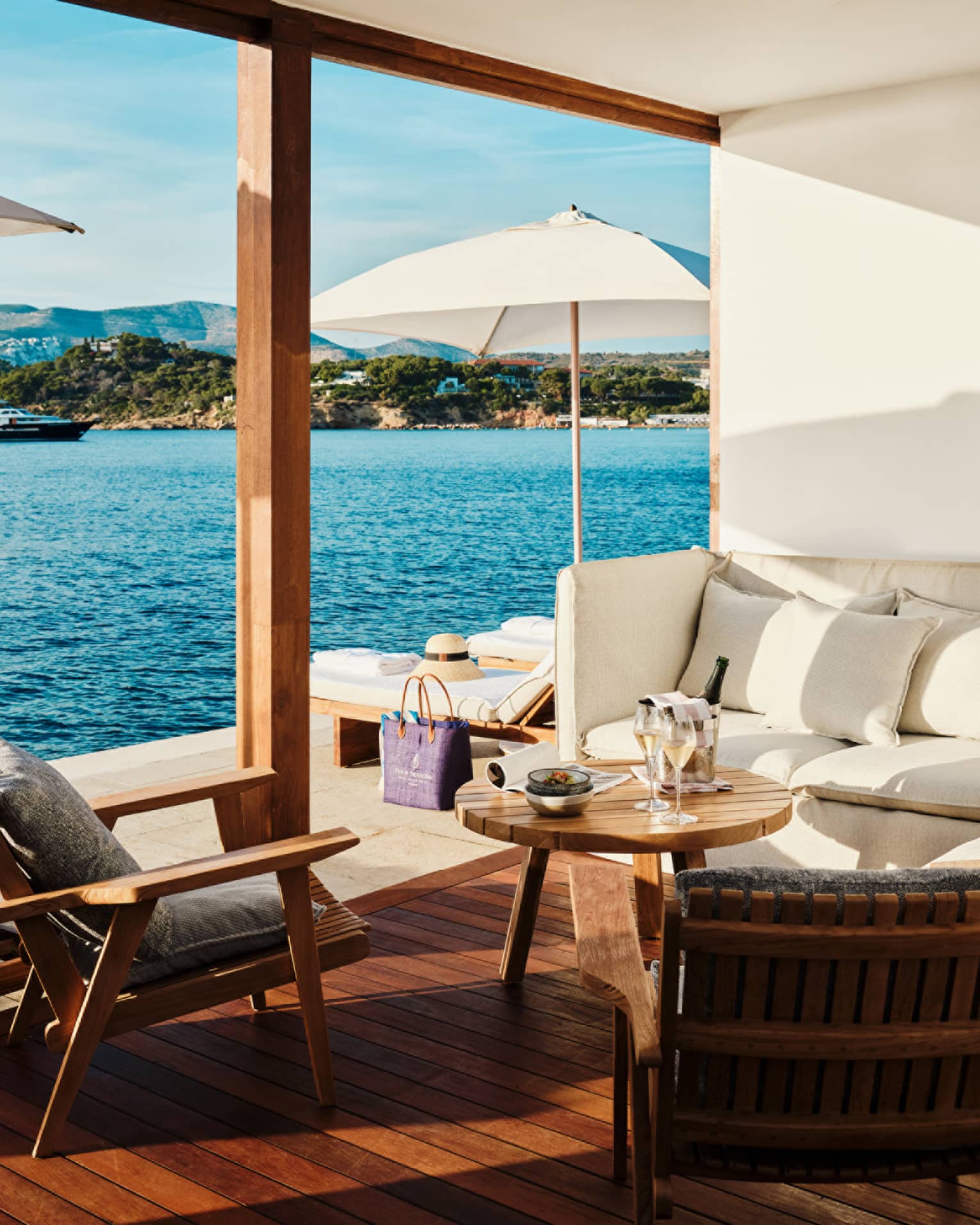 Cabana interior with white banquette-style couch, round teak table and two teak chairs open to platform with white umbrellas and blue sea views