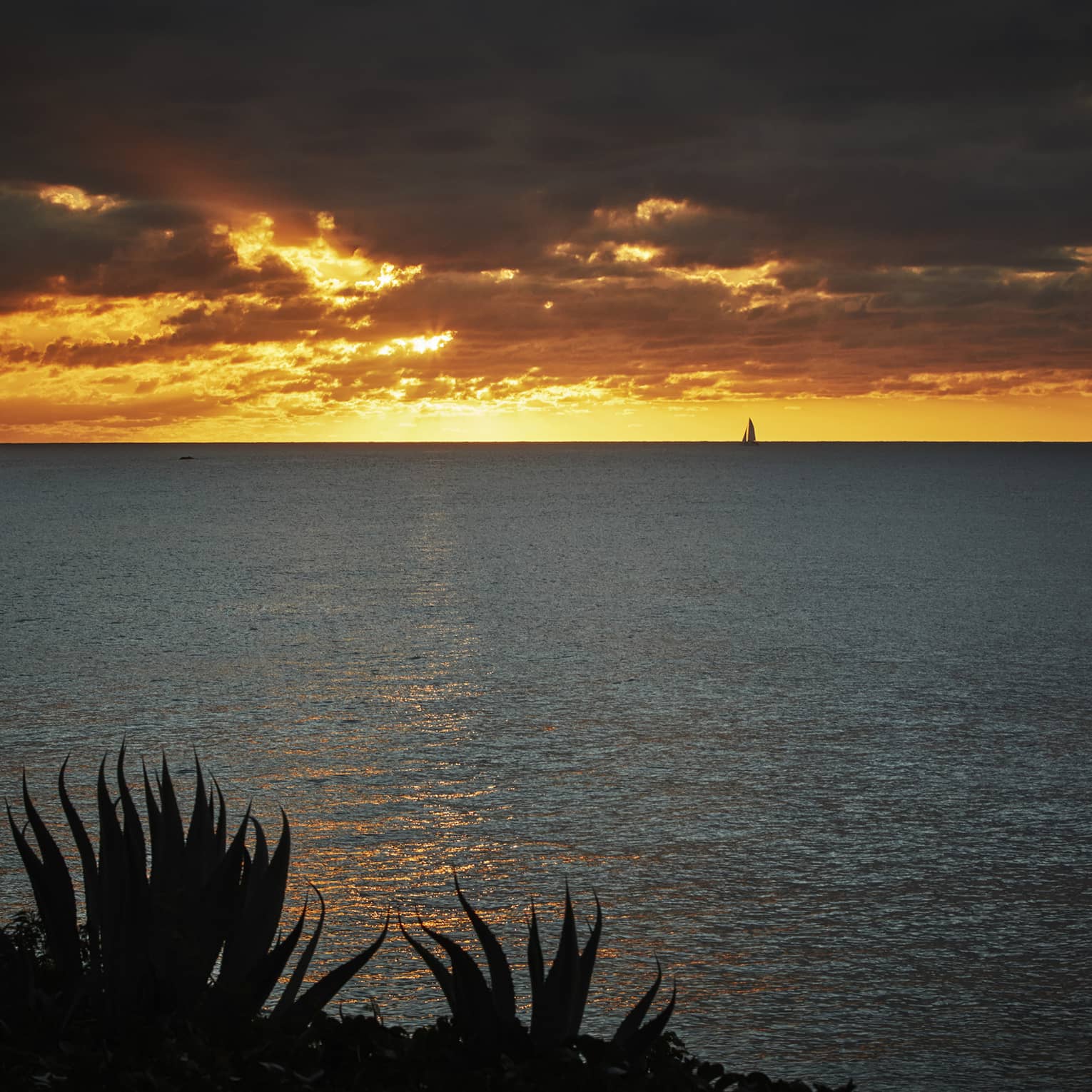 Bright orange sunset over the ocean with dramatic clouds and a sailboat in the distance