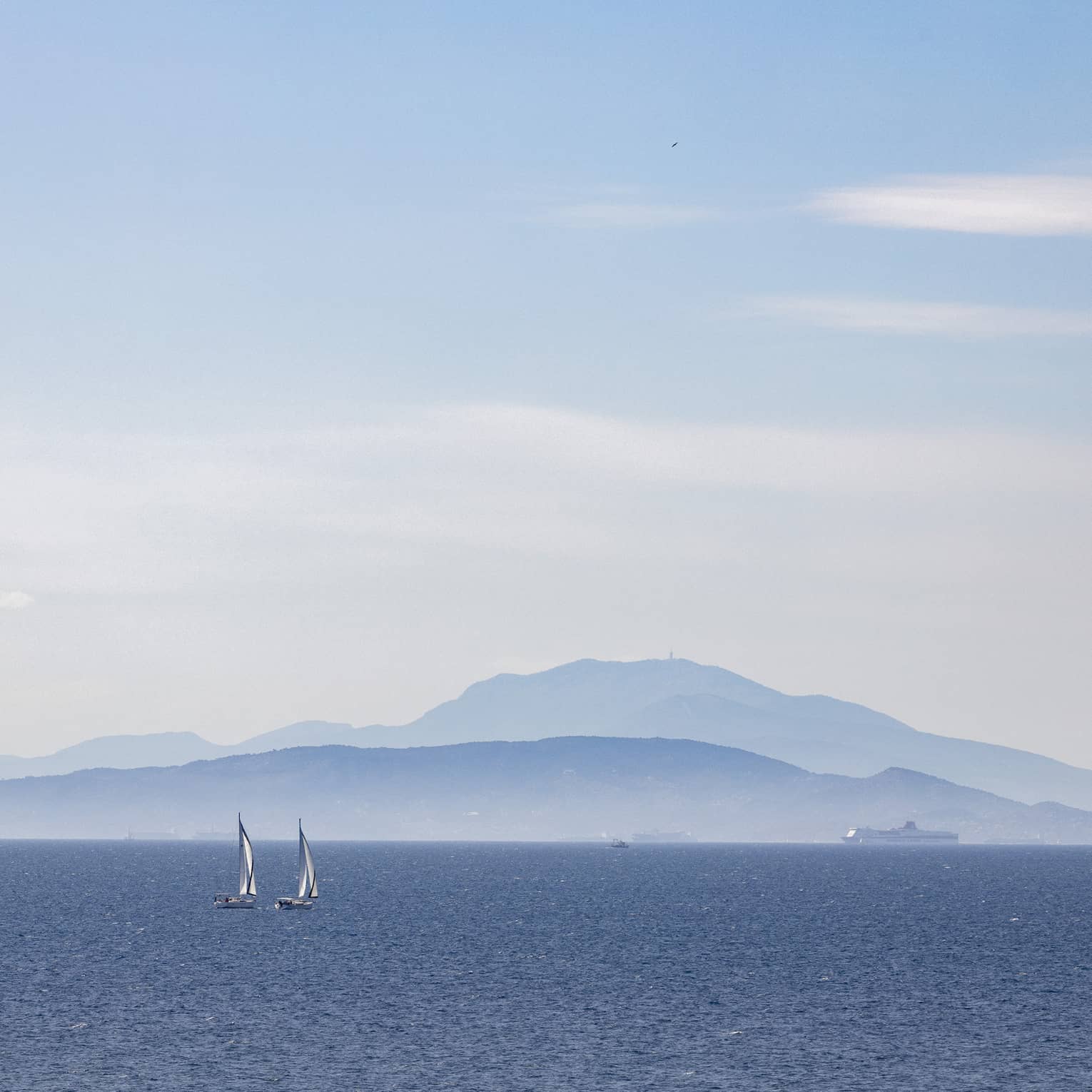 Two sailboats on blue ocean with blue-grey mountain range in the background 
