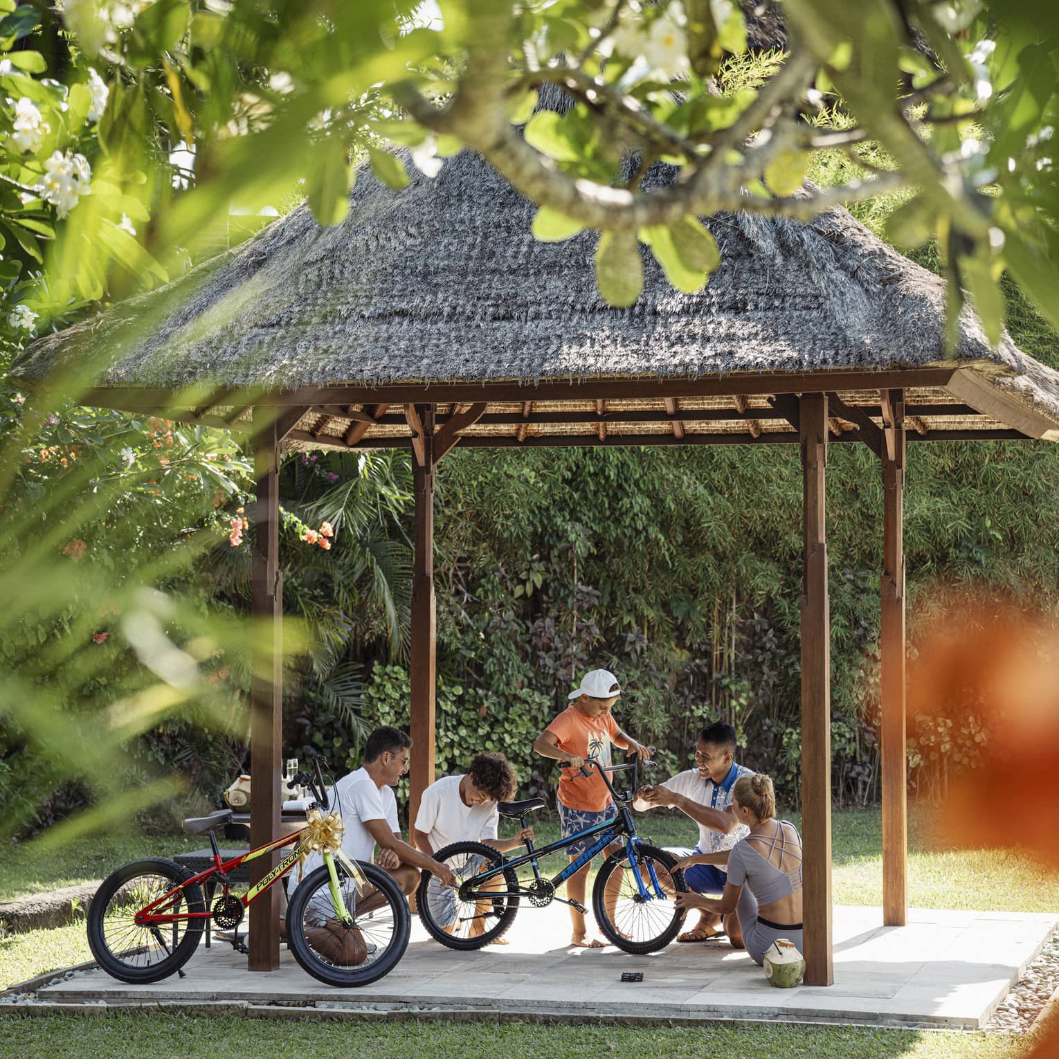 View through foliage of four adults and a child working on a bicycle under a thatched hut surrounded by lush greenery.