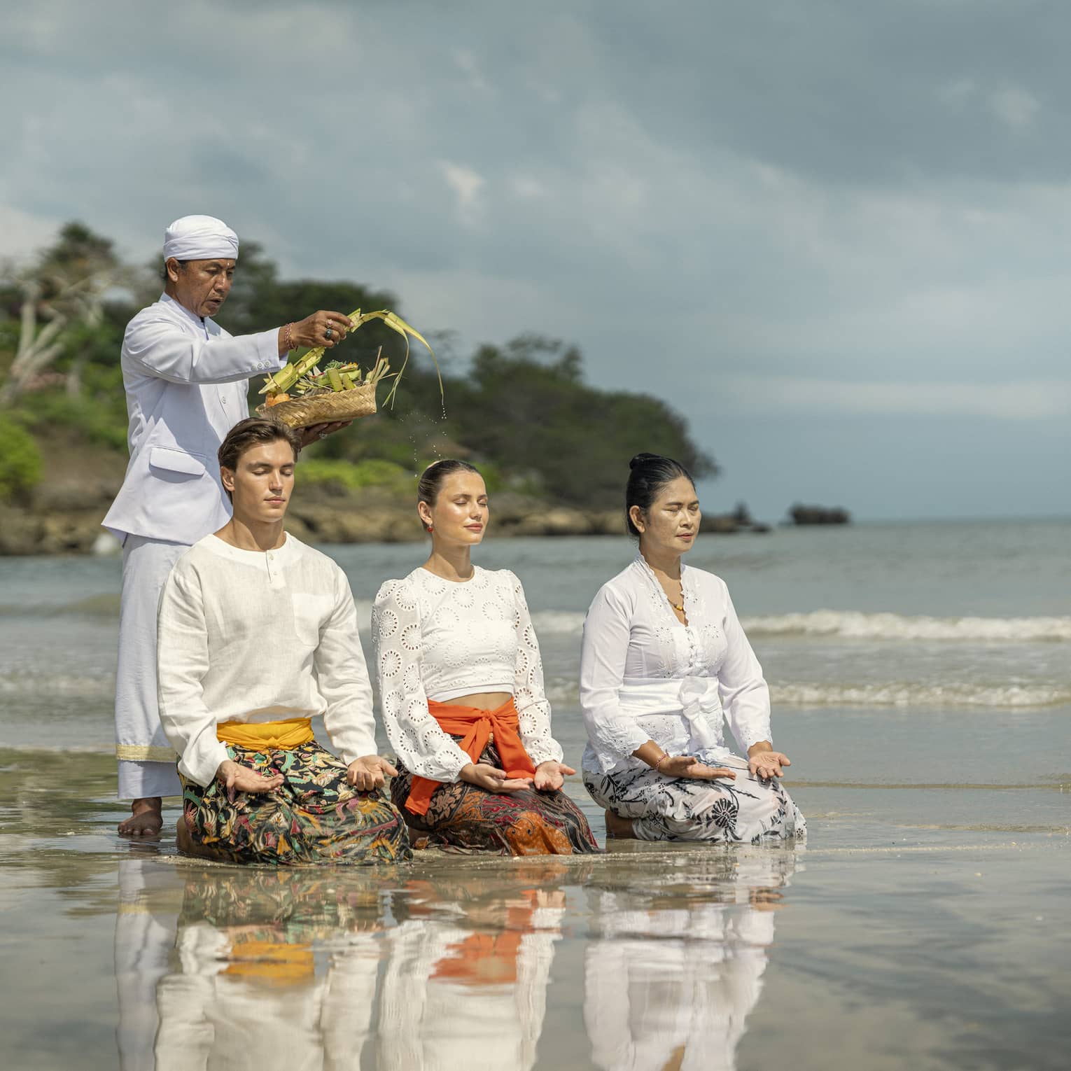 Three adults sit in shallow water with palms up on their knees as another sprinkles water from palm fronds on their heads.