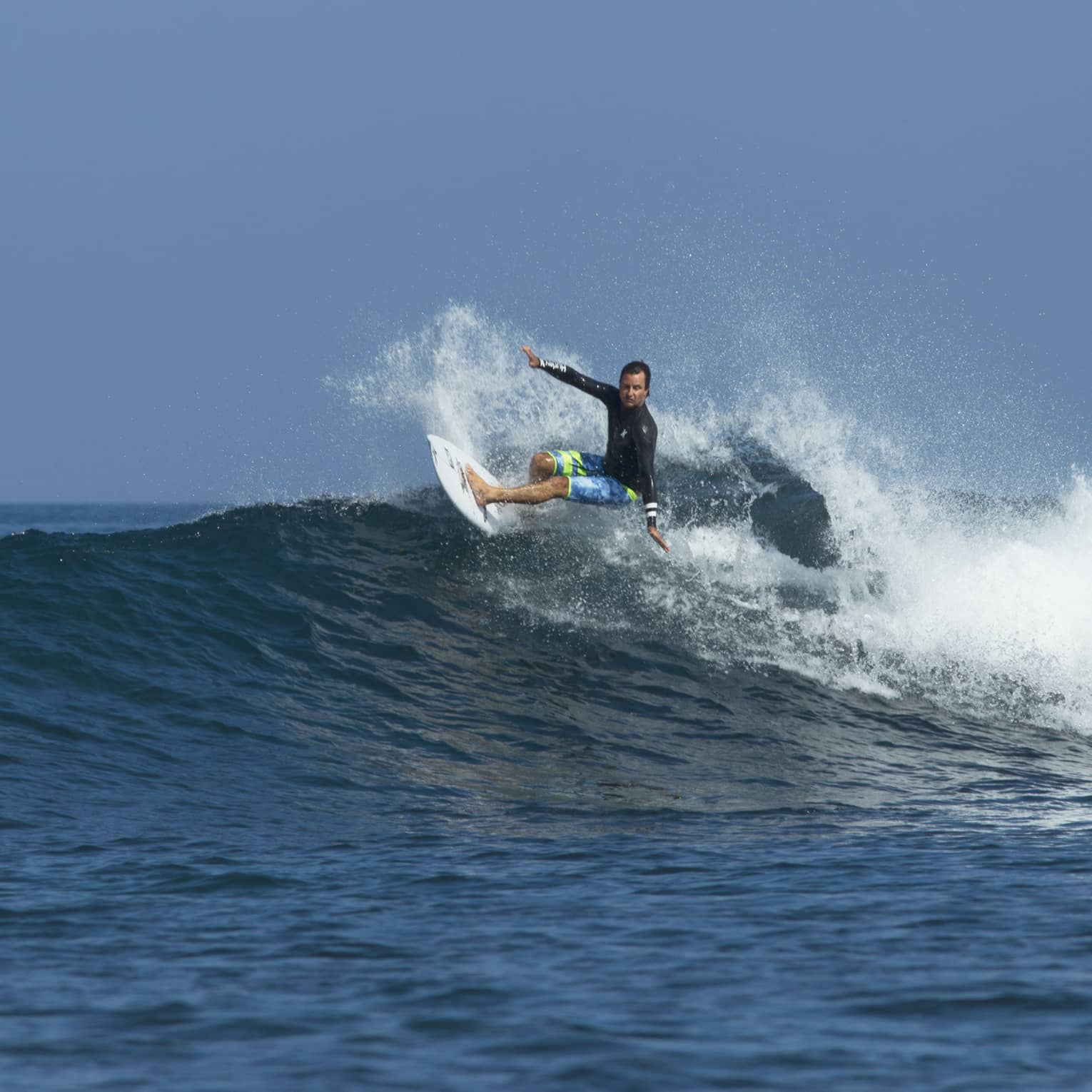 A surfer in a long sleeve rash guard and board shorts rides a wave, arms outstretched, in the wake of frothy white spray.