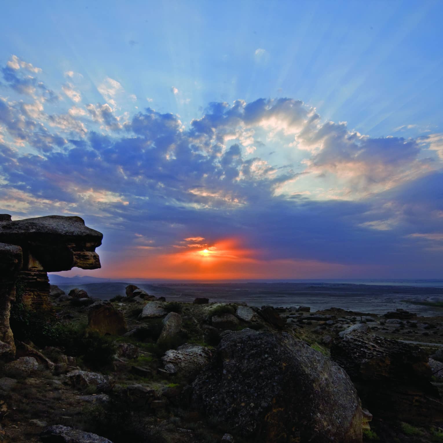 Sunbeams and fluffy azure blue clouds dance across a twilight sky, the vibrant red sun gently illuminating a rocky shoreline.