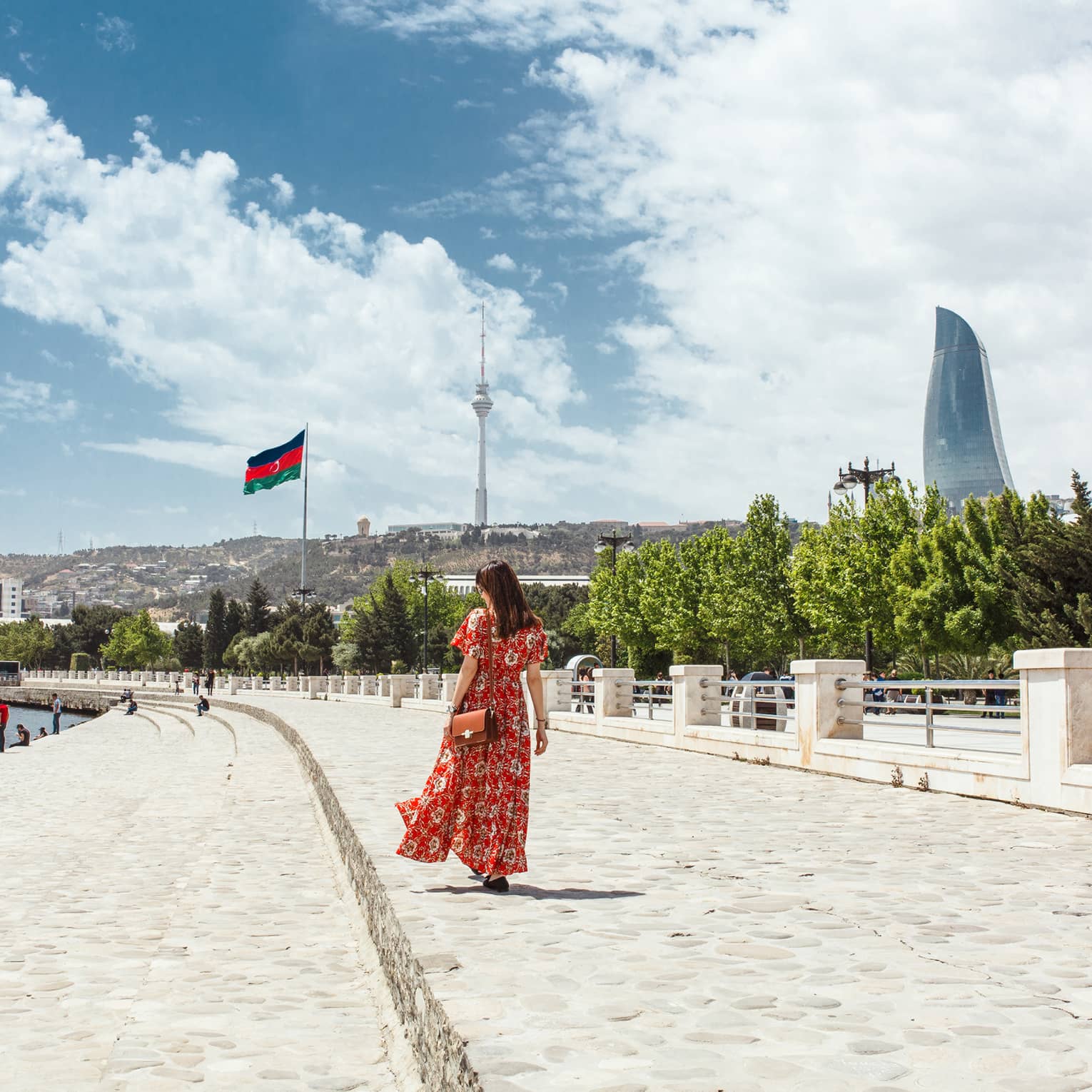 Woman wearing red dress walks along stone waterfront, skyscrapers in background