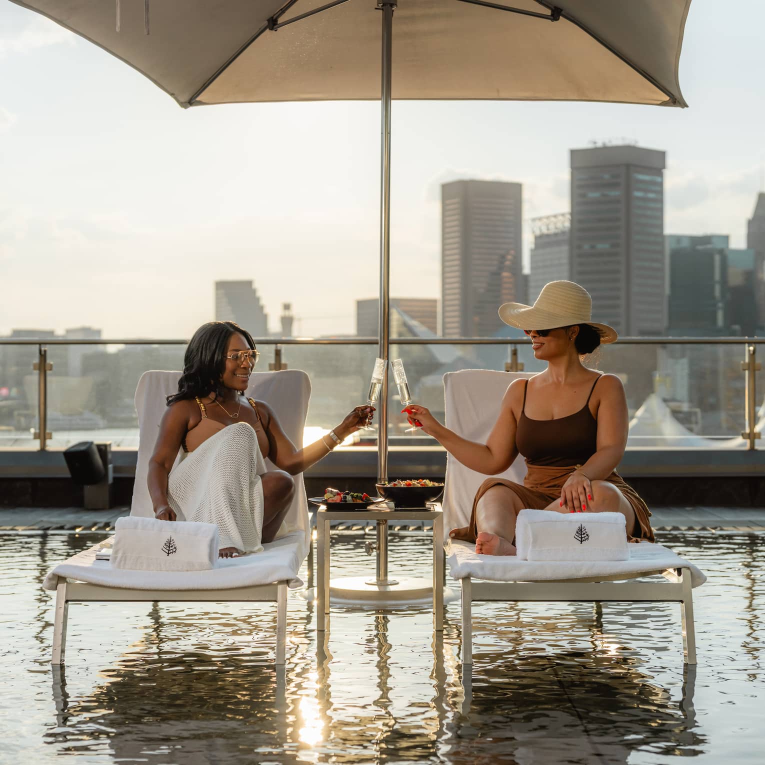 Two guests have a toast with drinks in stemmed glasses, while they sit on lounge chairs under a sun umbrella over a rooftop pool, with city skyline views in the background.