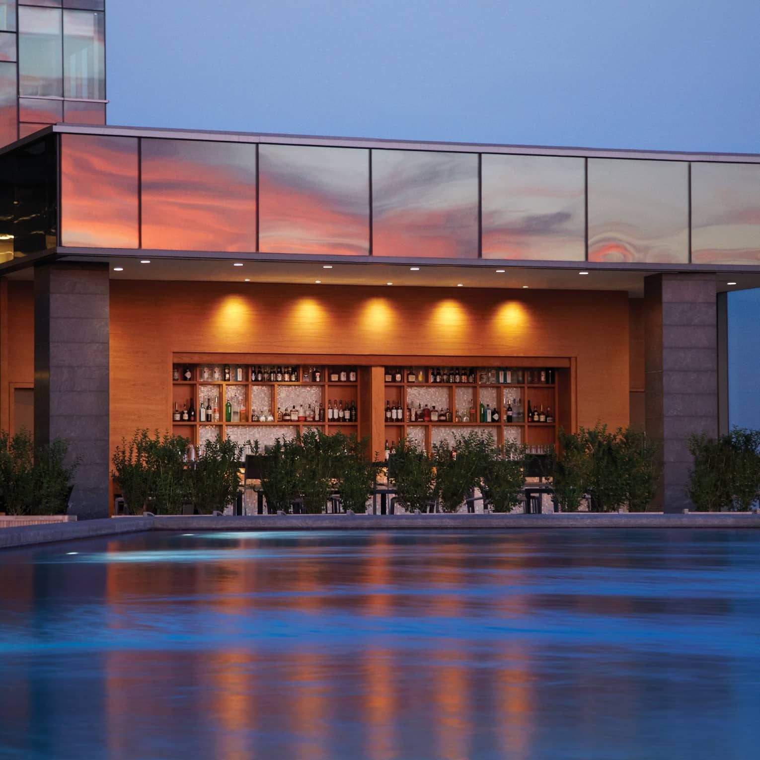 Illuminated swimming pool, wood bar and liquor displays under glass building exterior at sunset
