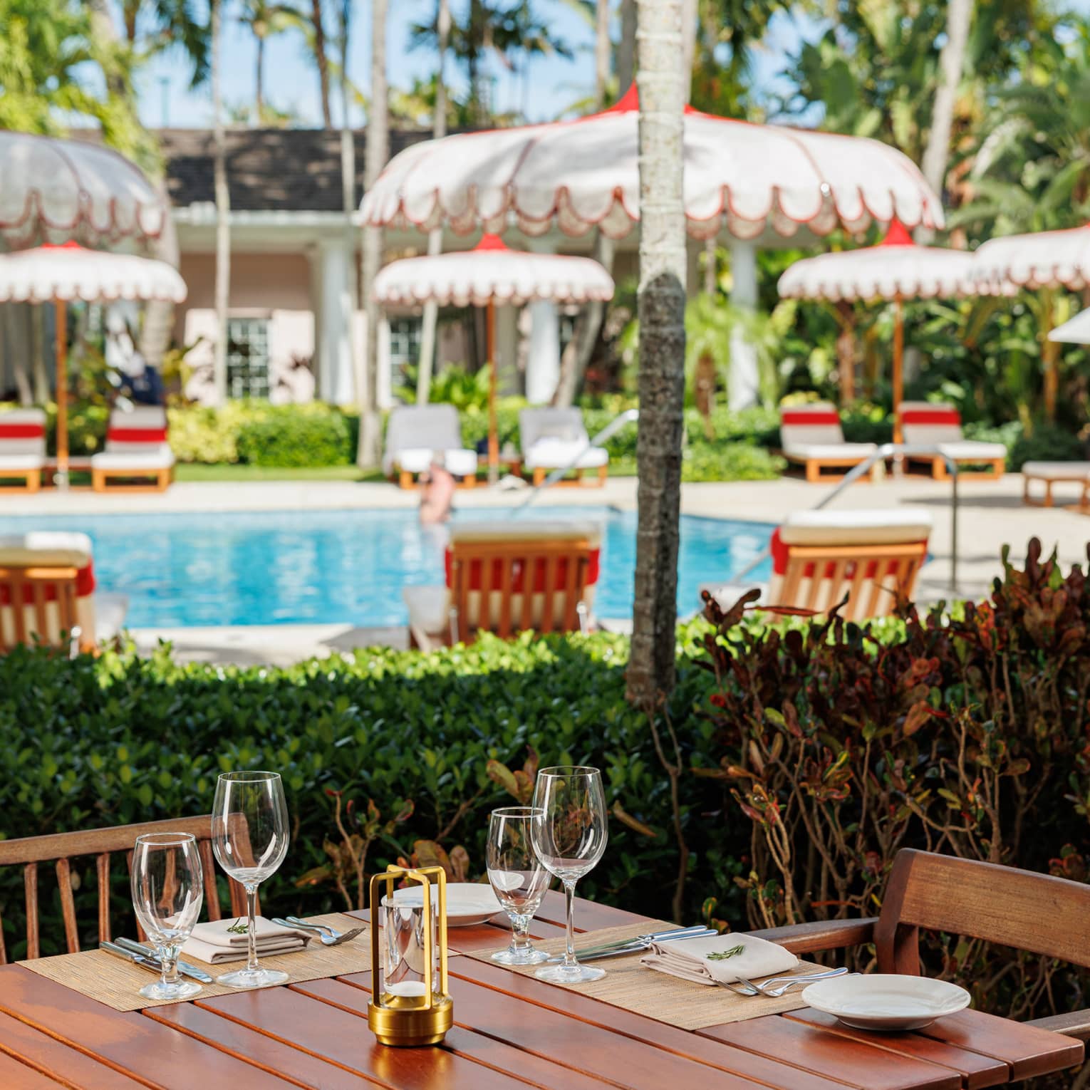 Wooden dining table topped with cutlery and glasses in front of an outdoor pool surrounded by white scalloped umbrellas and lounge chairs