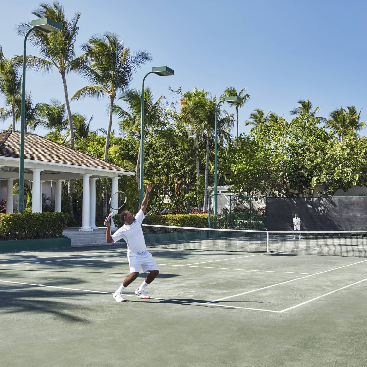 A man in all white trows a tennis ball high into the air, mid-serve 