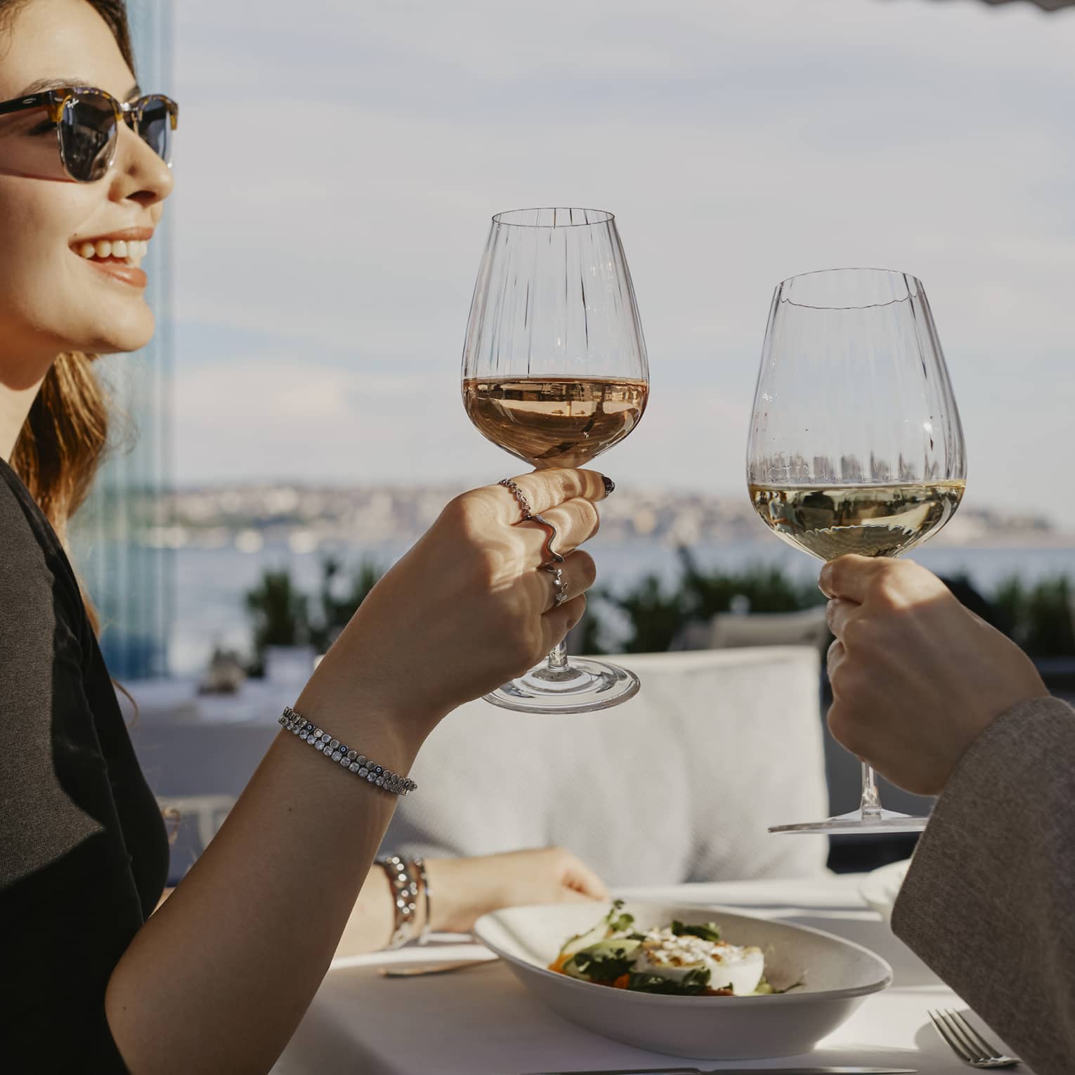 Person wearing sunglasses, smiling while toasting with a glass of wine with another person
