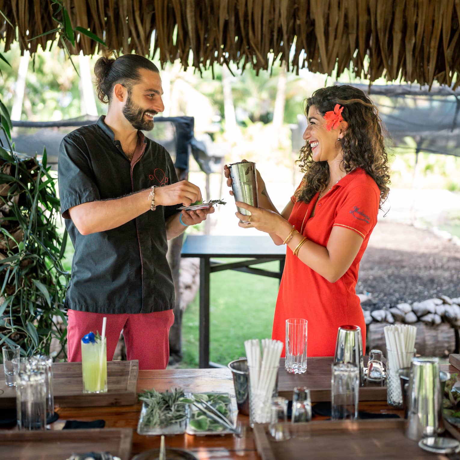 Two people hold a shaker and fresh herbs in a straw-roofed hut over a bar strewn with glasses, liquor bottles and garnishes.