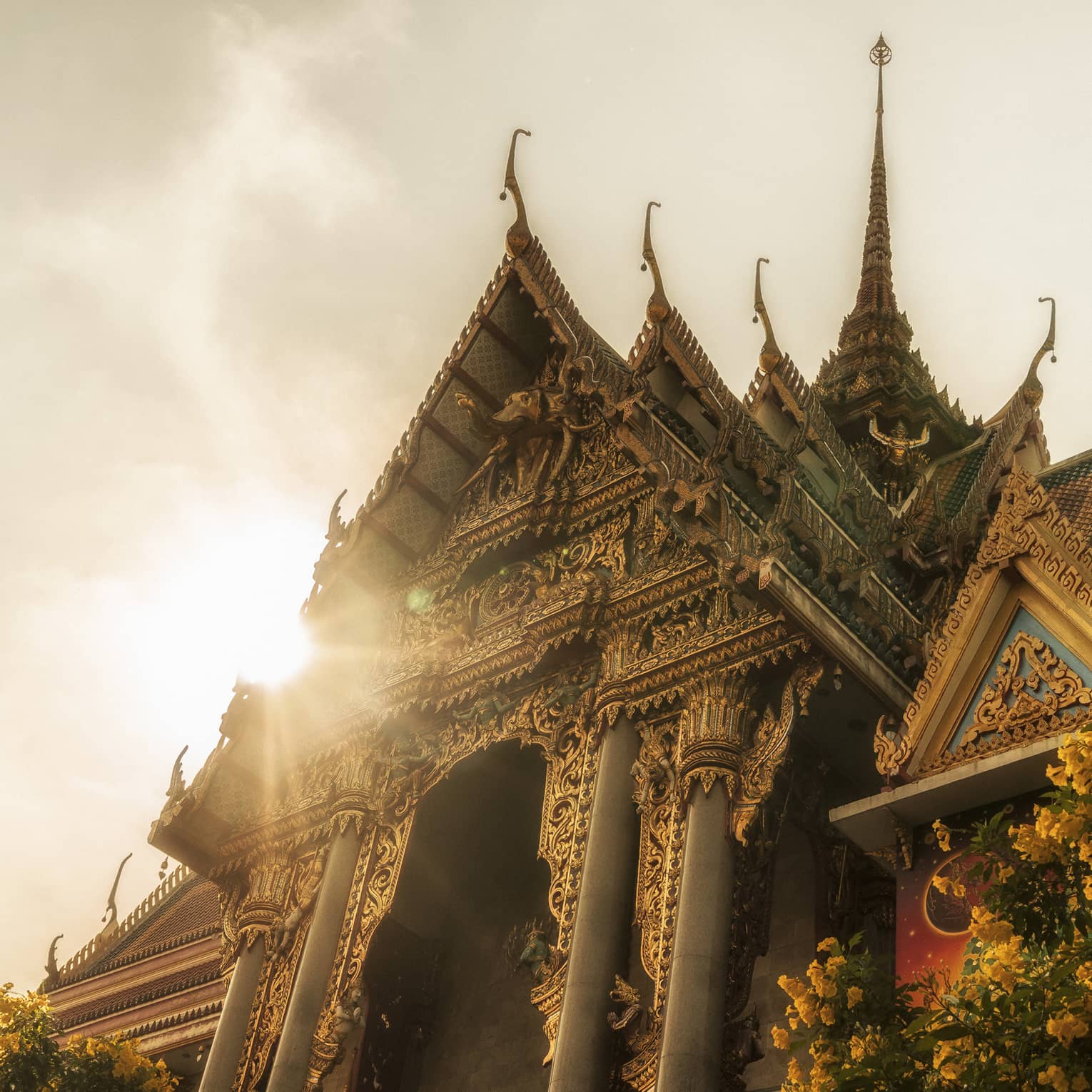 Intricately designed temple with ornate carvings and tiered roofs, illuminated by sunlight, with a tree and yellow flowers in the foreground
