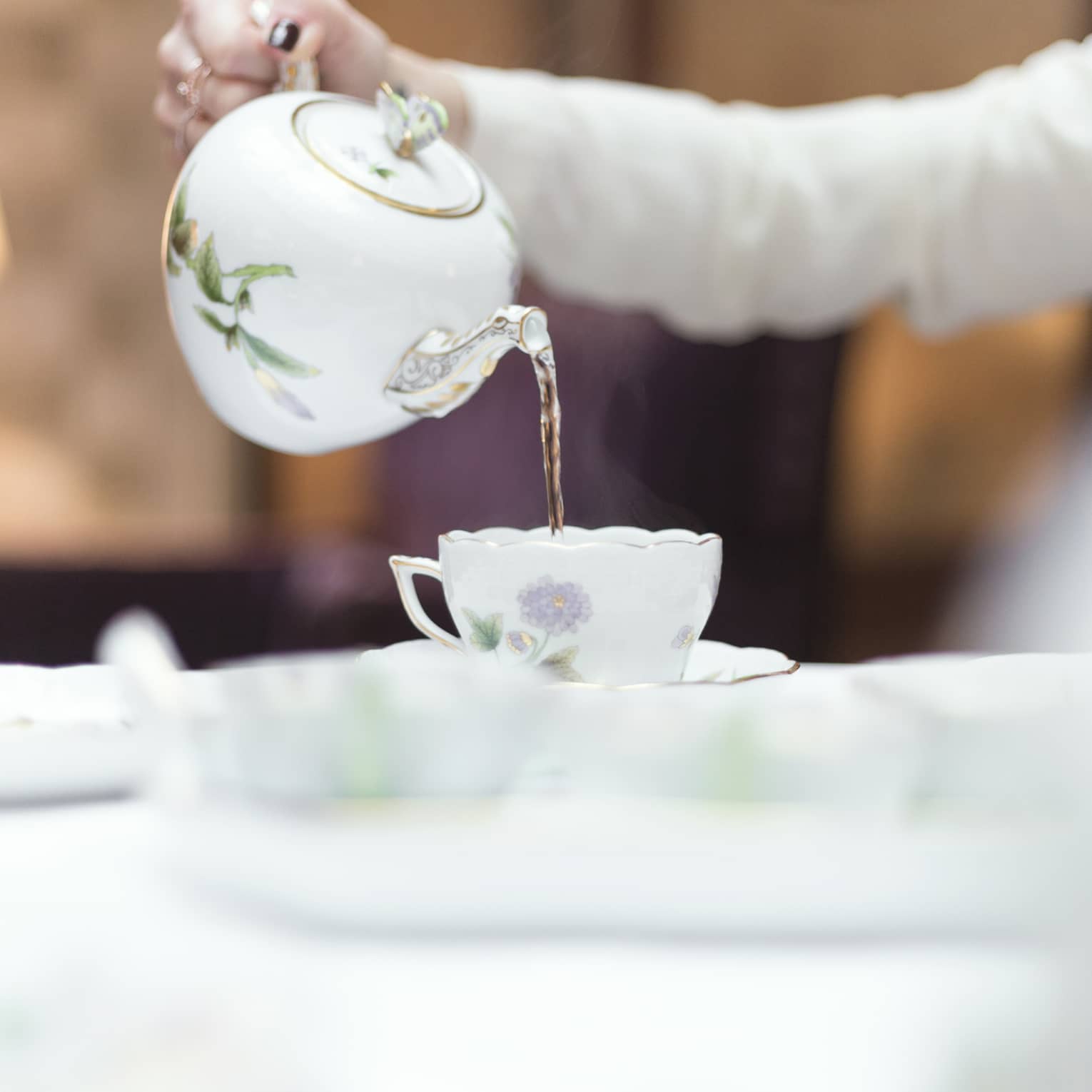 Person holds floral tea pot and pours tea into cup and saucer 