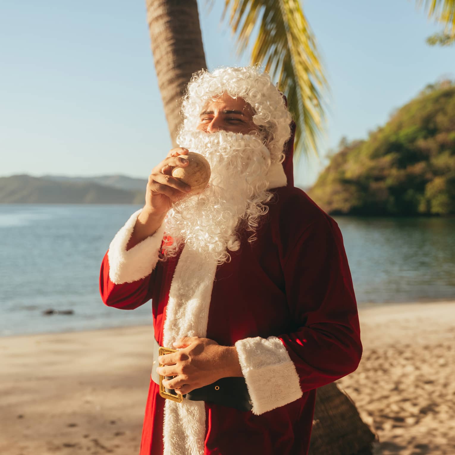 A person on the beach wearing a Santa Claus costume and holding a coconut