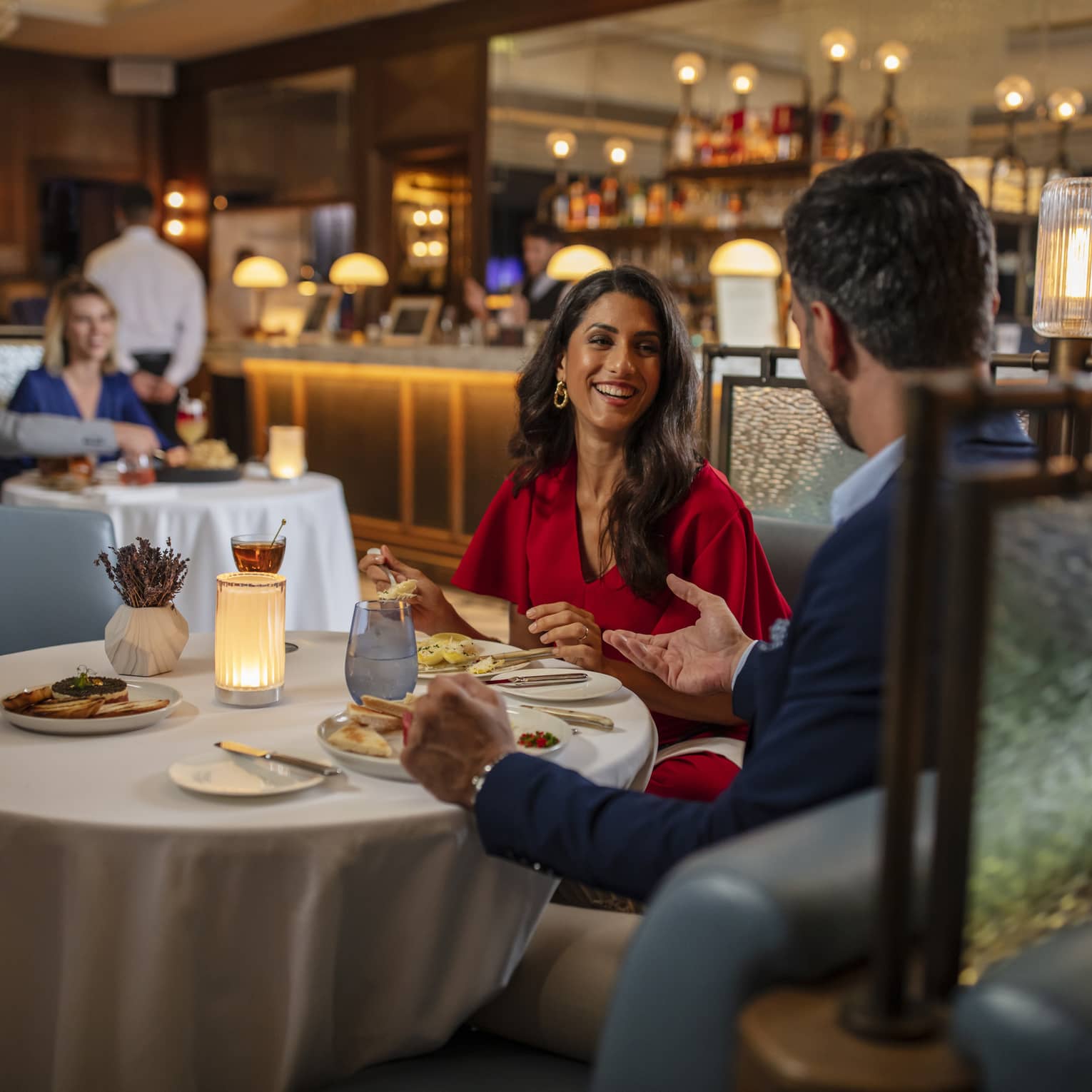 Woman in red dress, smiling at man, seated at round dining table in bar lounge