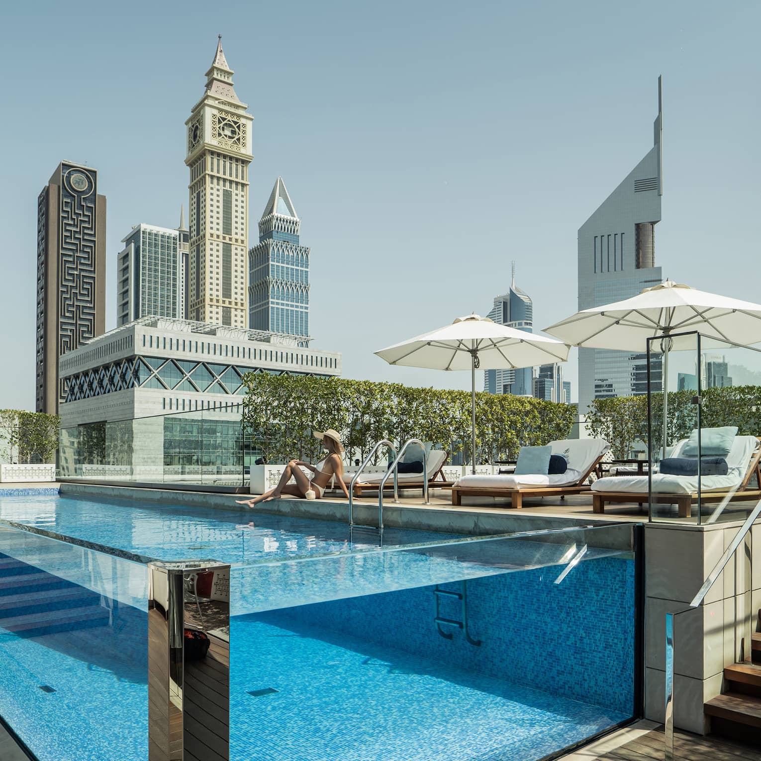 A guest lounges by a transparent rectangular rooftop pool, modern towers with geometric designs rising into the azure sky.