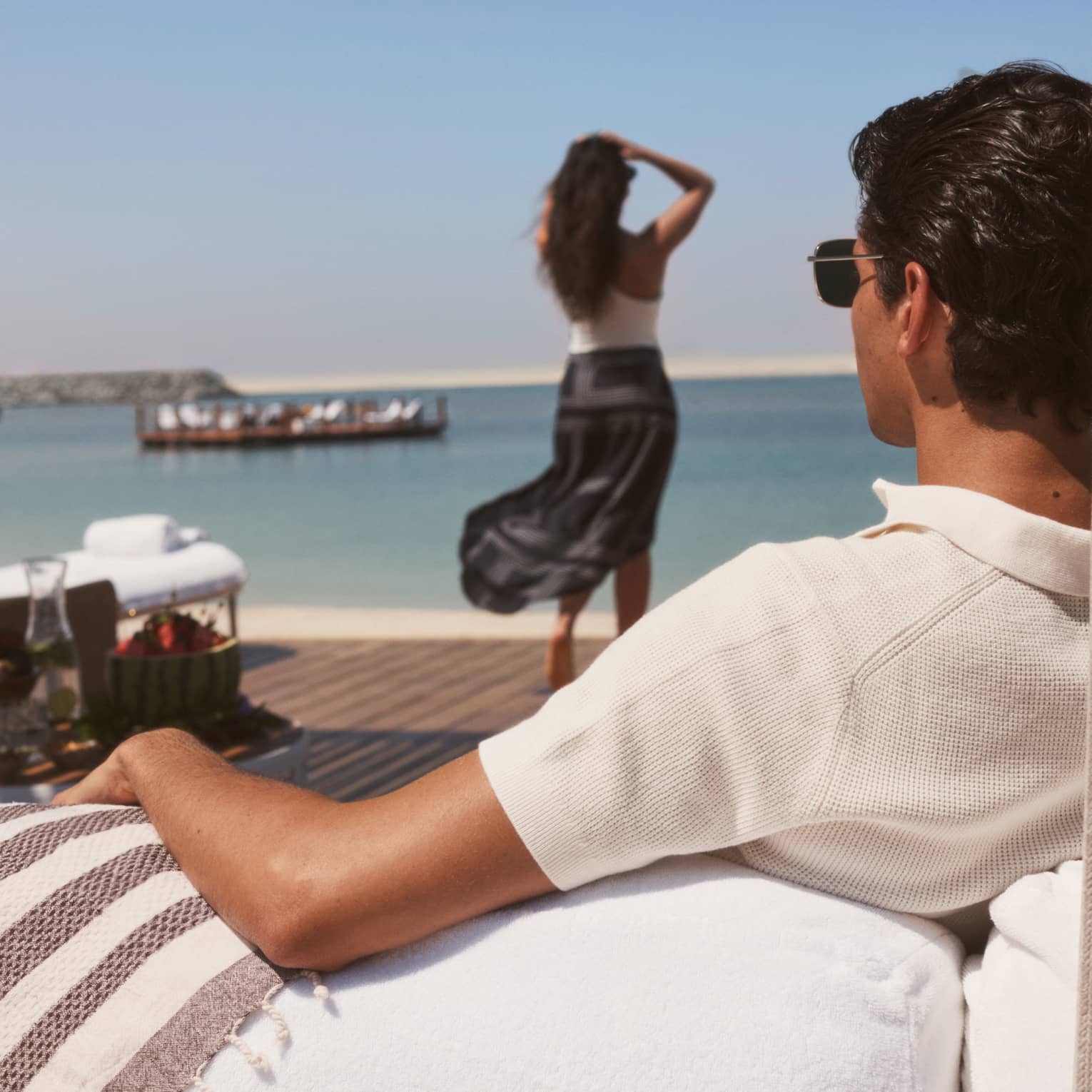 Man wearing white polo shirt lounges in a beachfront cabana while a woman wearing a white top and black maxi skirt walks toward the water