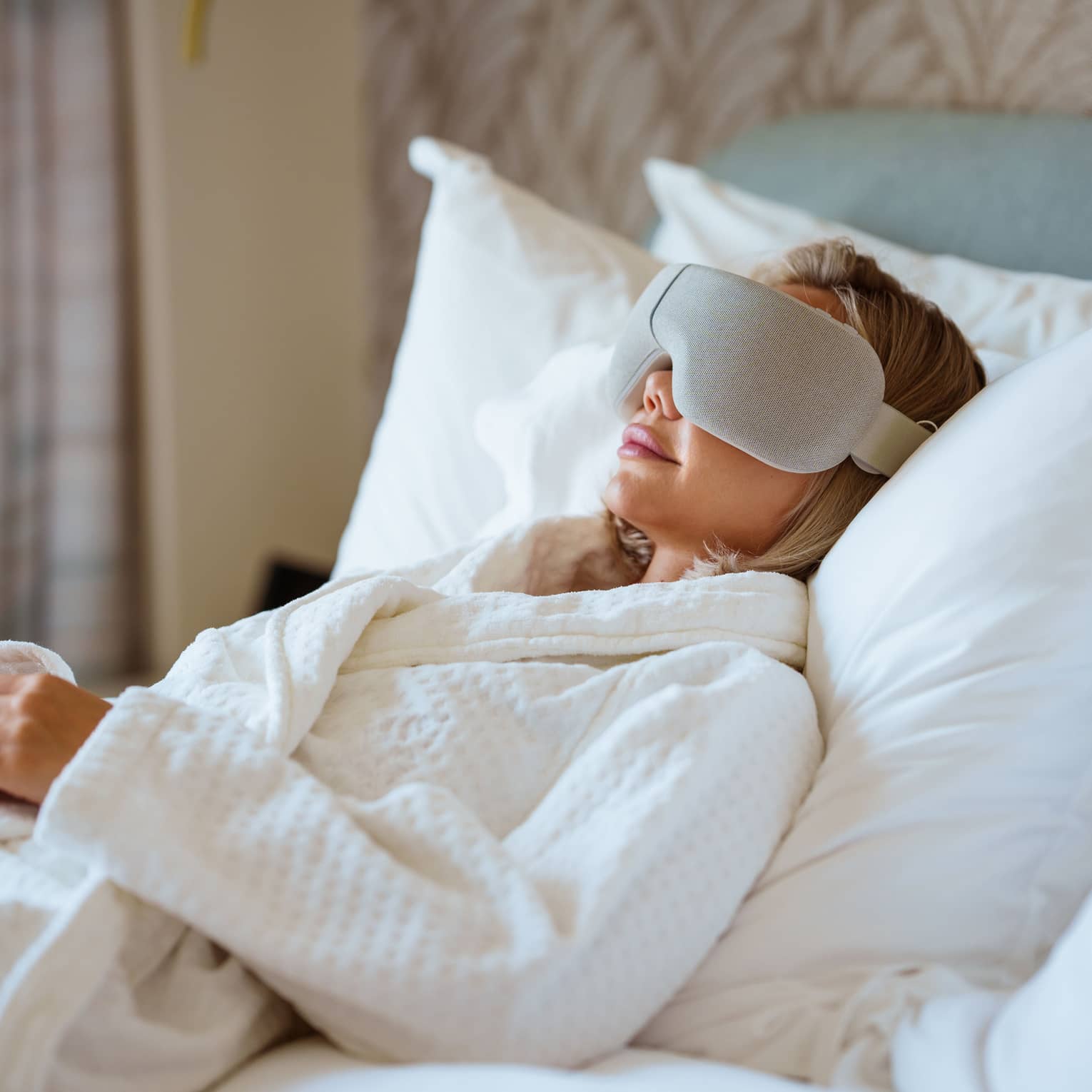 Person wearing white spa robe lays back on a bed with large white pillows wearing a high-tech eye mask