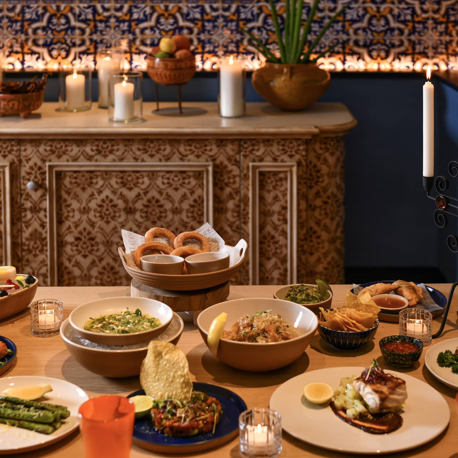 A table set with a variety of appetizers on white plates and bowls with a sideboard in the background adorned with white pillar candles