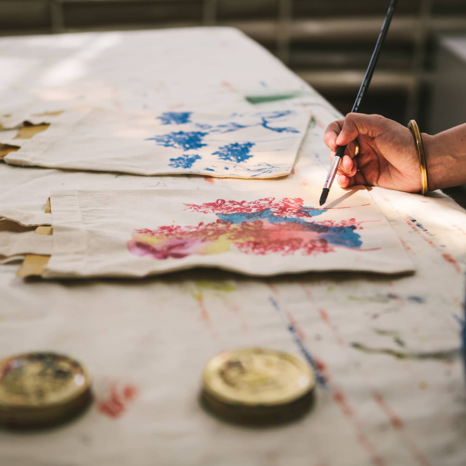 Close-up of a gold-bangled hand wielding a long brush to paint an intricate pattern on a cloth tote bag.